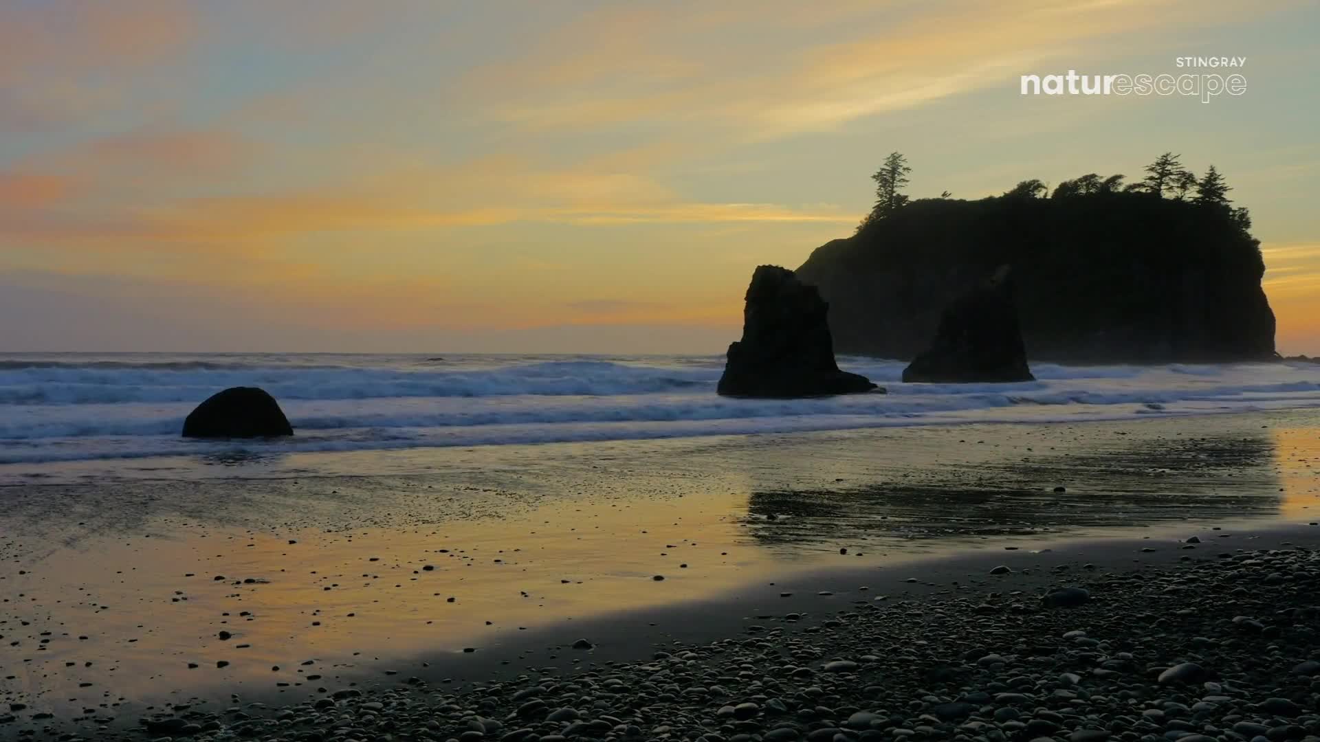 Waves crash against the rocky shore as the sun dips below the horizon. The sky glows with soft oranges and yellows, reflecting on the wet sand.