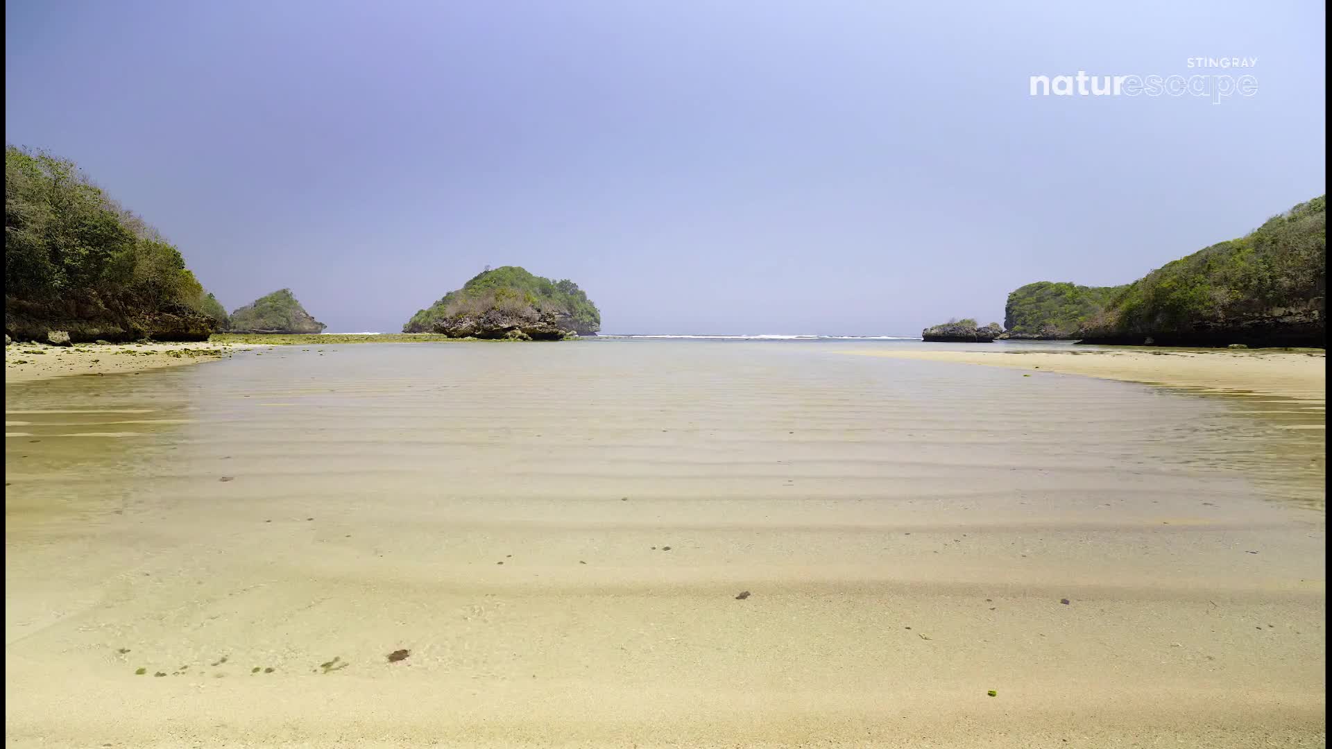 The shallow water ripples across the sand, revealing a seabed etched with delicate lines.  Green, tree-covered hills rise from the water's edge on either side, framing a calm horizon.