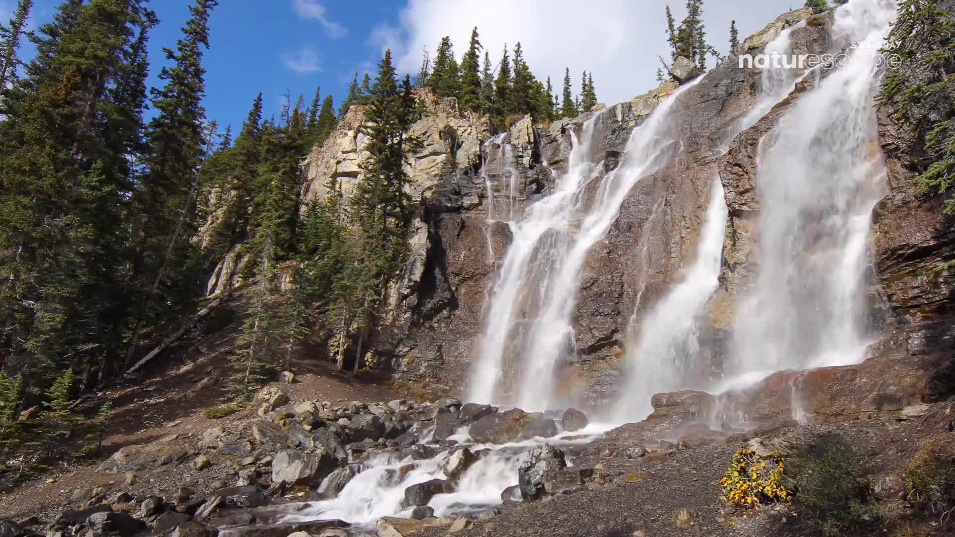 Water cascades down a rocky Canadian mountainside, fanning out into a mist at its base. A faint rainbow arcs through the spray.