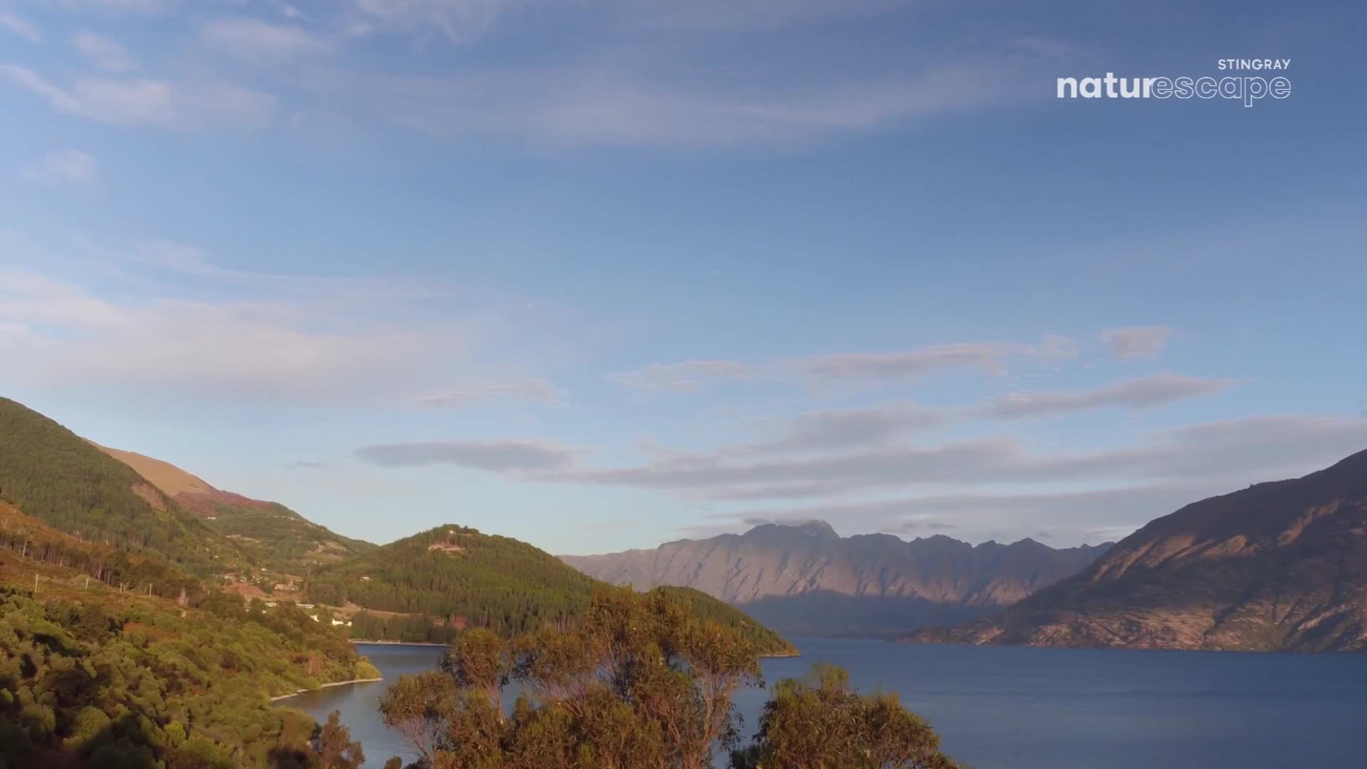 The sun casts long shadows across the forested hills that slope down to a wide, calm lake. Jagged mountains rise in the distance, their peaks catching the late afternoon light.