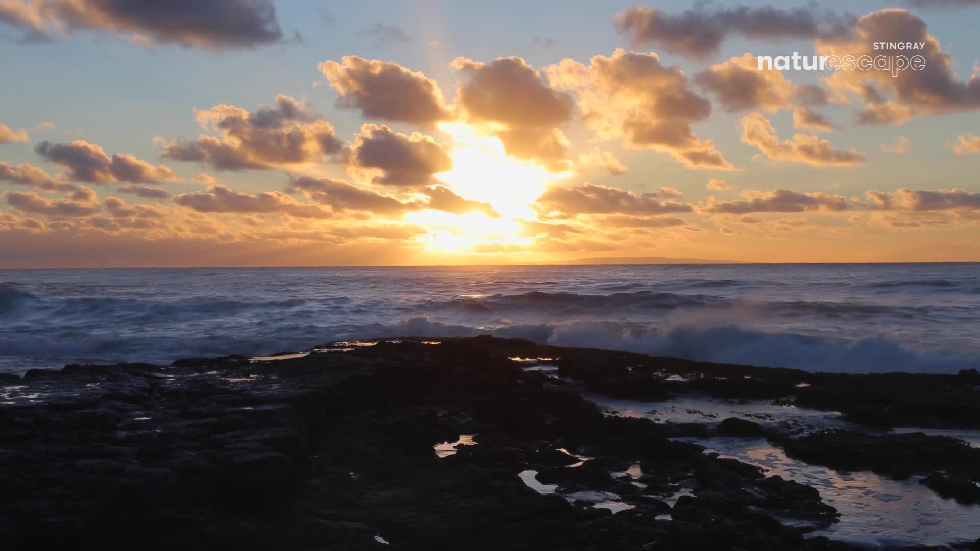 The sun breaks through the clouds, casting a golden glow on the restless ocean. Waves crash against the dark, wet rocks in front of me, reflecting the sky's fiery colours.