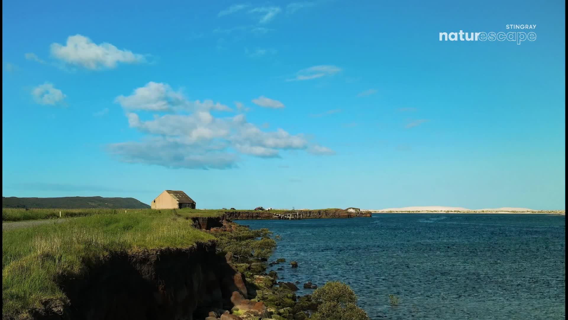 The water laps against the rocky shore under a vast Canadian sky. A small, weathered building sits atop the grassy cliff overlooking the bay.