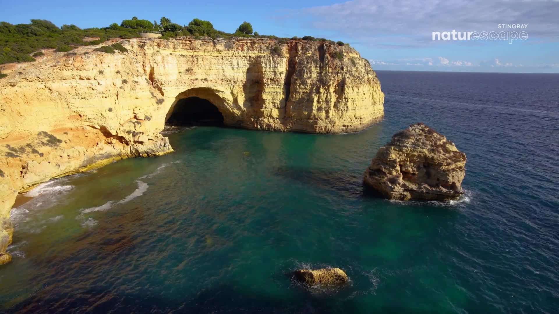 The turquoise water laps at the base of a massive, sun-baked cliff, revealing a dark cave opening. A jagged rock formation stands sentinel in the deep blue sea.
