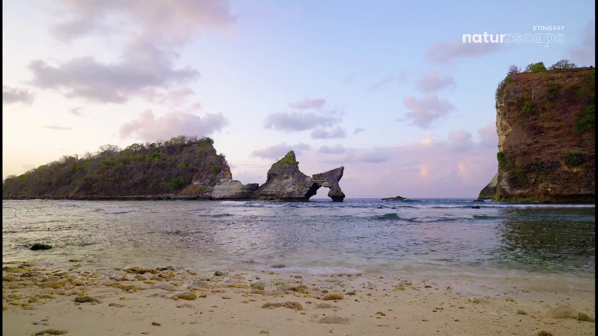 Gentle waves lap the sandy shore, revealing a natural arch carved into the rock formations. The sky above glows with soft hues of pink and purple as clouds drift lazily.