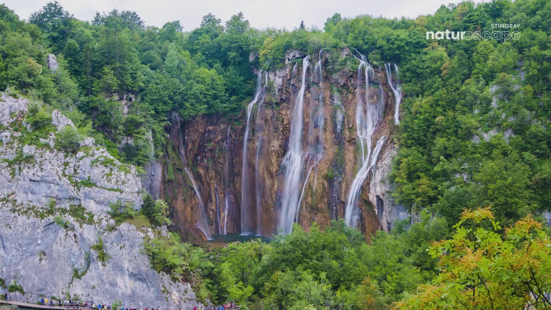 Water cascades down a massive, moss-covered cliff face, a sight that rivals any Canadian natural wonder. A wooden boardwalk snakes along the base, crowded with people admiring the scene.