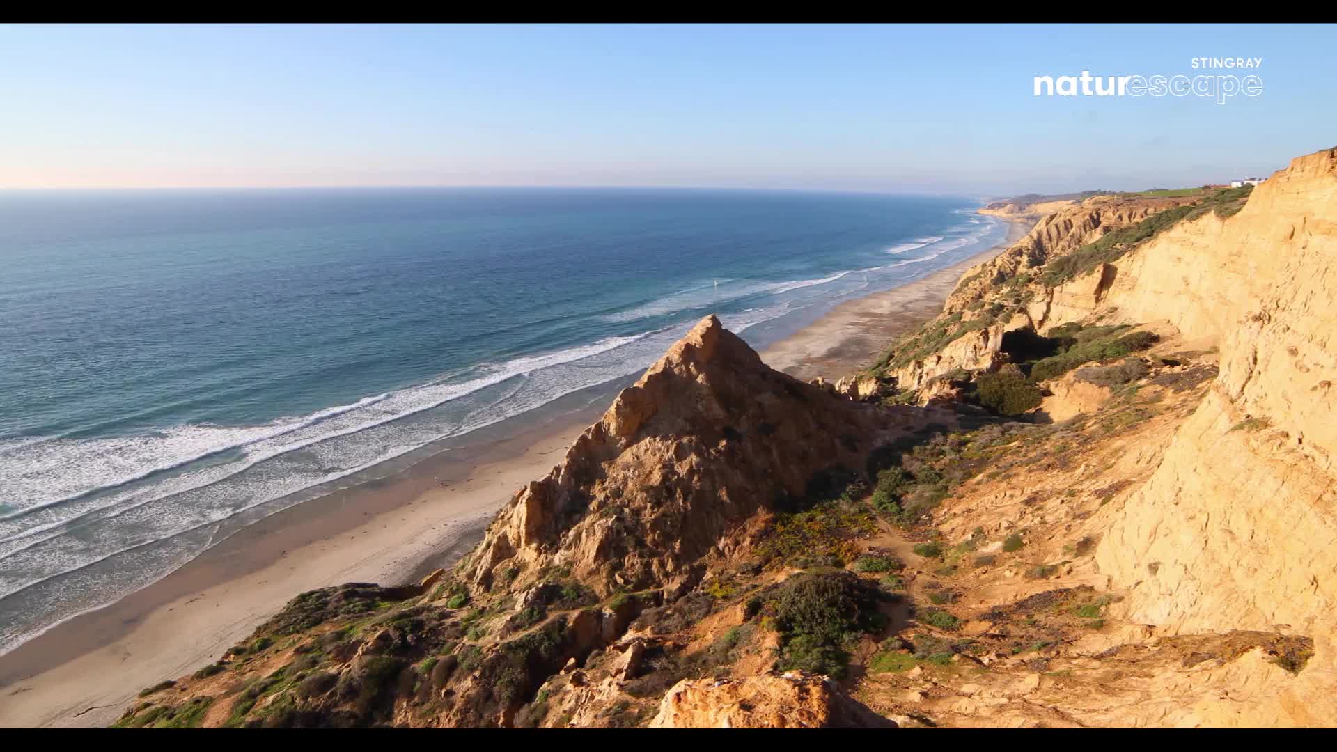 Waves roll onto a wide, sandy beach stretching along the coast. The ocean's deep blue meets the pale sky, with rugged, sun-baked cliffs rising sharply to my right.