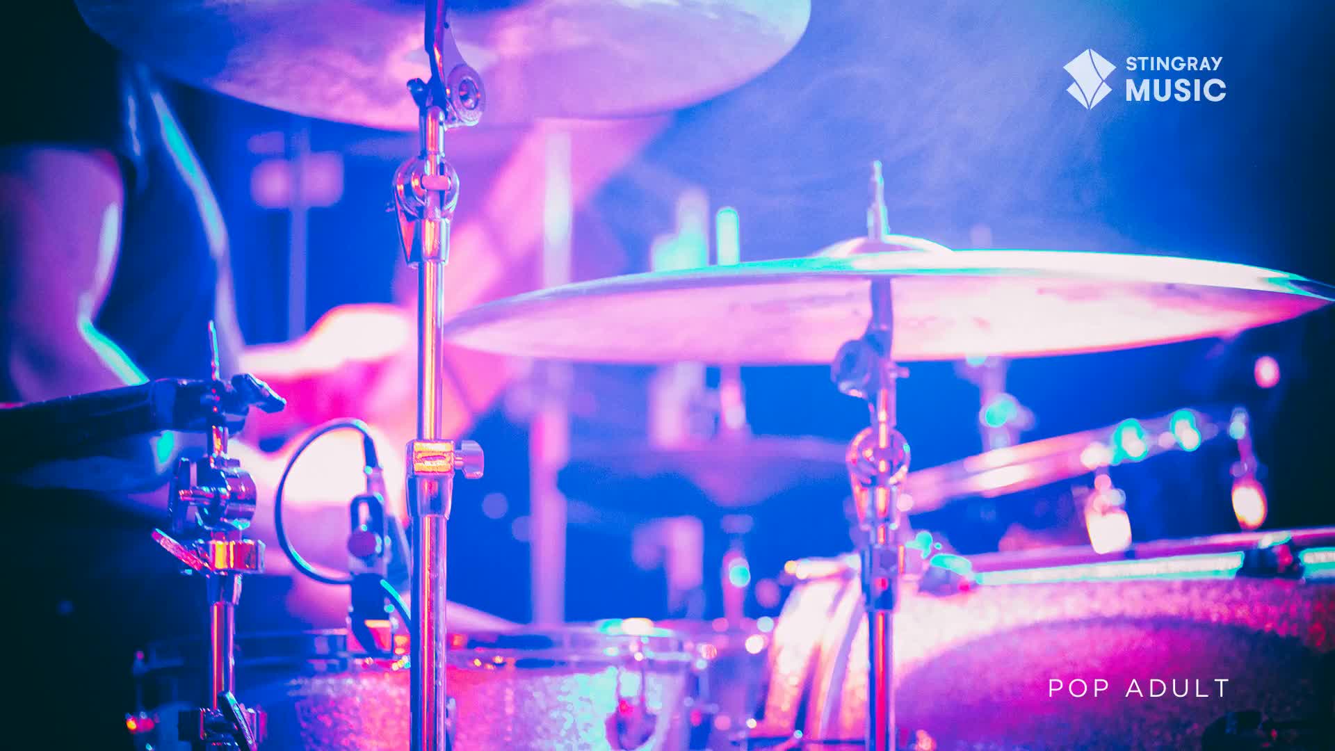 The drummer's arms are a blur, striking the cymbals and snare with a steady rhythm. The stage lights reflect off the drum kit, casting a purple and blue glow over the scene.
