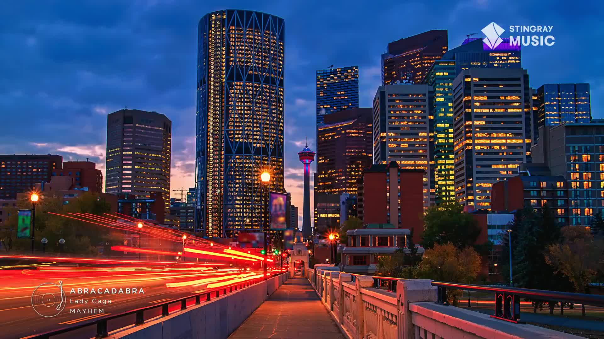 The city lights are on, and streaks of red and orange from passing vehicles paint the bridge. Towering buildings, lit up with countless windows, stand tall against the twilight sky, and the Calgary Tower is lit up in the distance.
