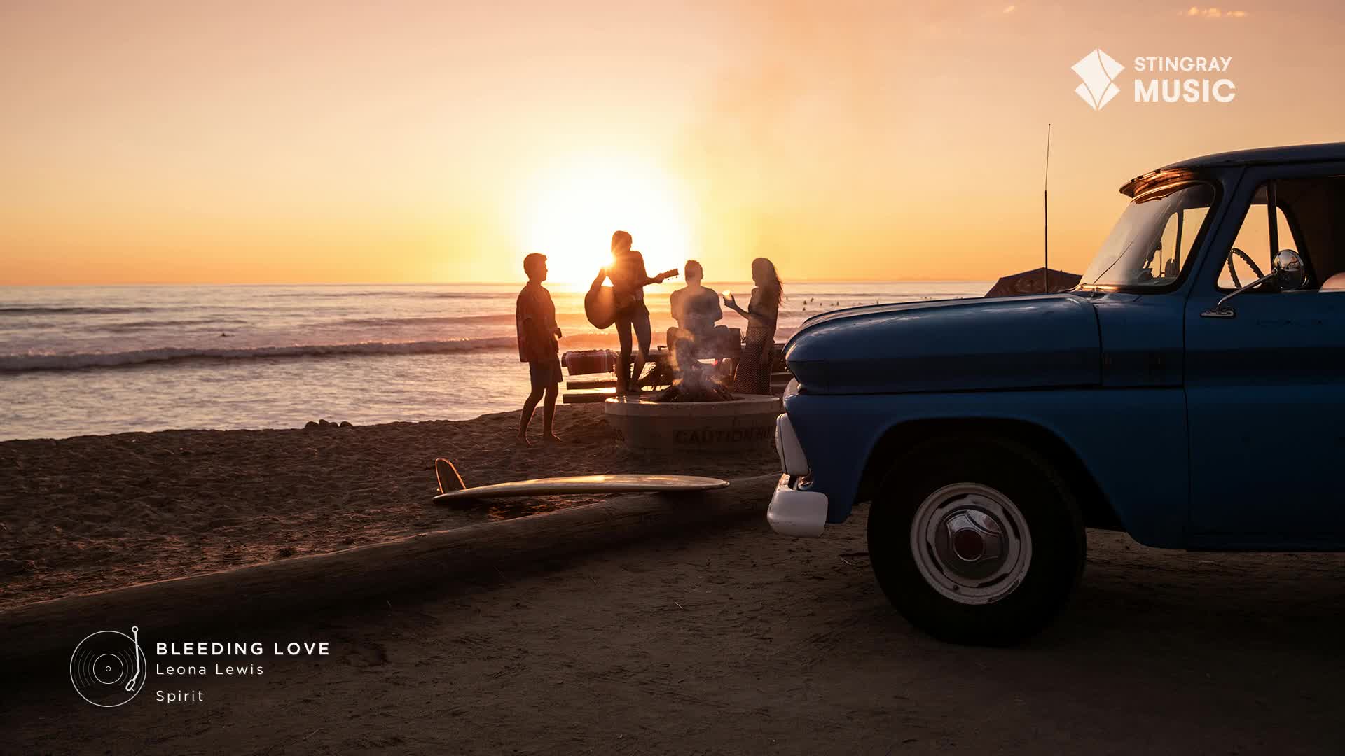 A band plays on a concrete platform near the beach, silhouetted against a sunset. The scene is framed by the front of a blue pickup truck and a surfboard lying on the sand.
