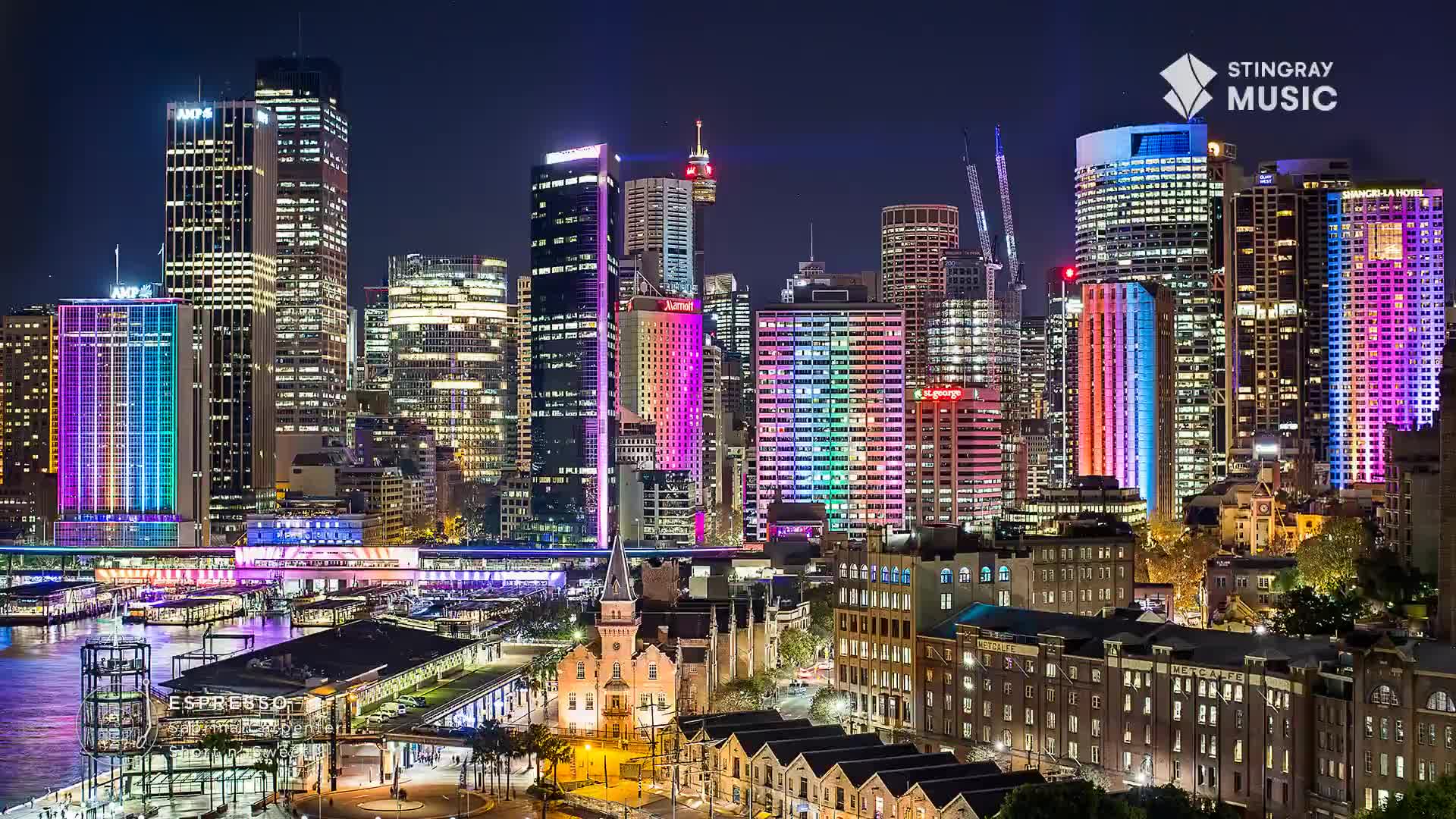 The city skyline glows, illuminated by vibrant lights, with the Stingray Music logo visible in the upper right. Buildings are awash in rainbow hues, reflecting off the water below.

