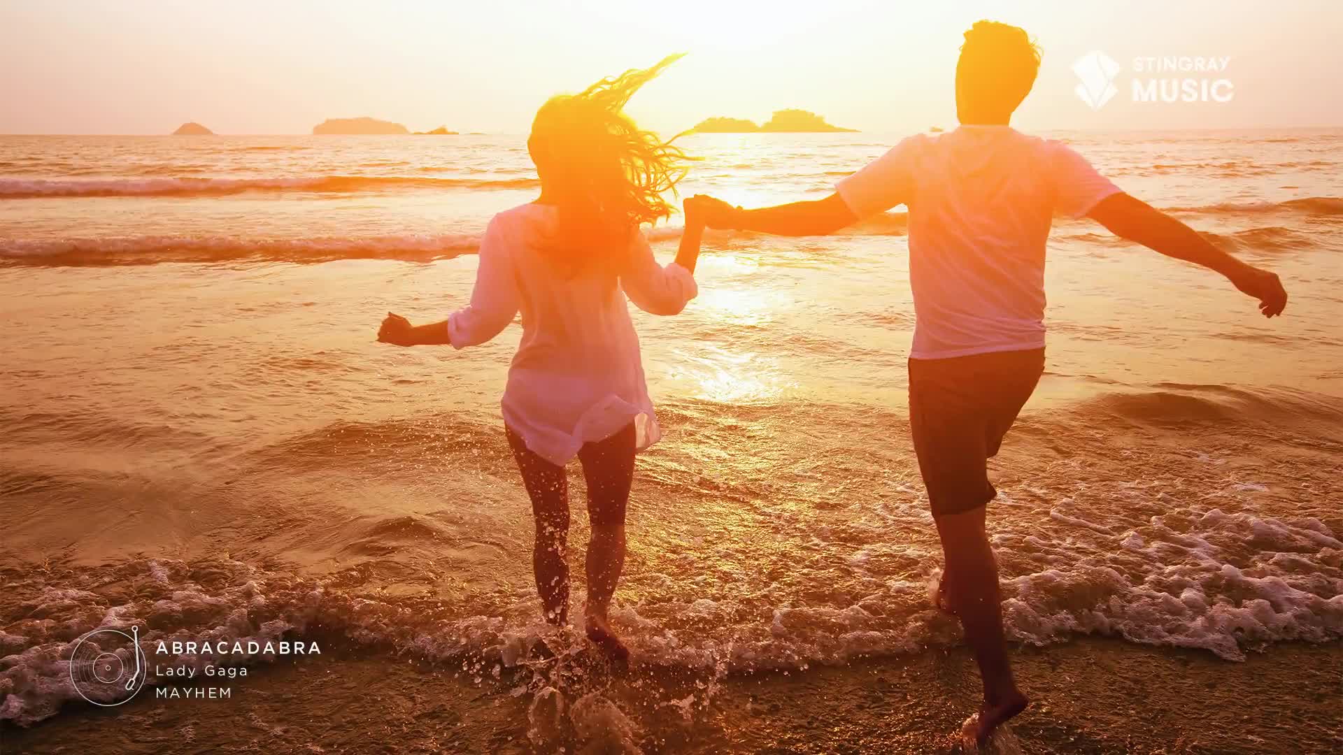 A couple, hand-in-hand, runs into the surf, their silhouettes outlined by the setting sun. The woman's hair whips back, and the man's arms are outstretched as they splash through the water.
