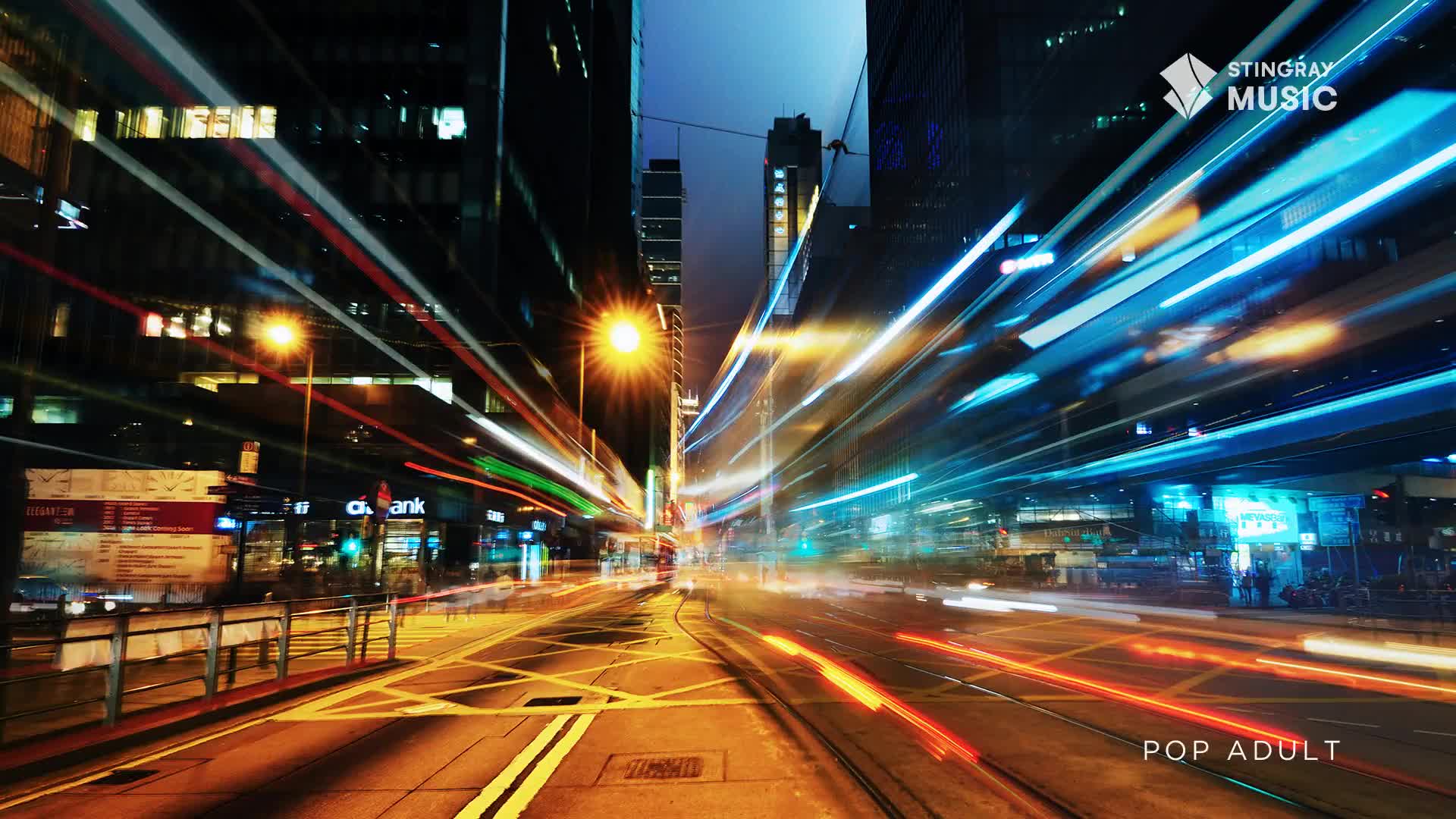 Bright streaks of light, likely from vehicles, race down a city street, creating long trails of color. The scene is framed by tall buildings, and the Stingray Music logo is visible in the upper right.
