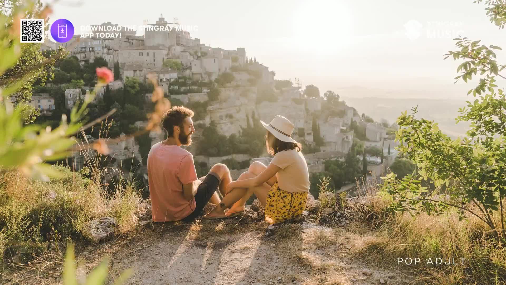 A couple sits close together, legs crossed, on a rocky outcrop overlooking a sun-drenched town. The man, in a pink shirt, looks at the woman in a hat and yellow skirt, their hands touching.
A couple sits close together, legs crossed, on a rocky outcrop overlooking a sun-drenched town. The man, in a pink shirt, looks at the woman in a hat and yellow skirt, their hands touching.