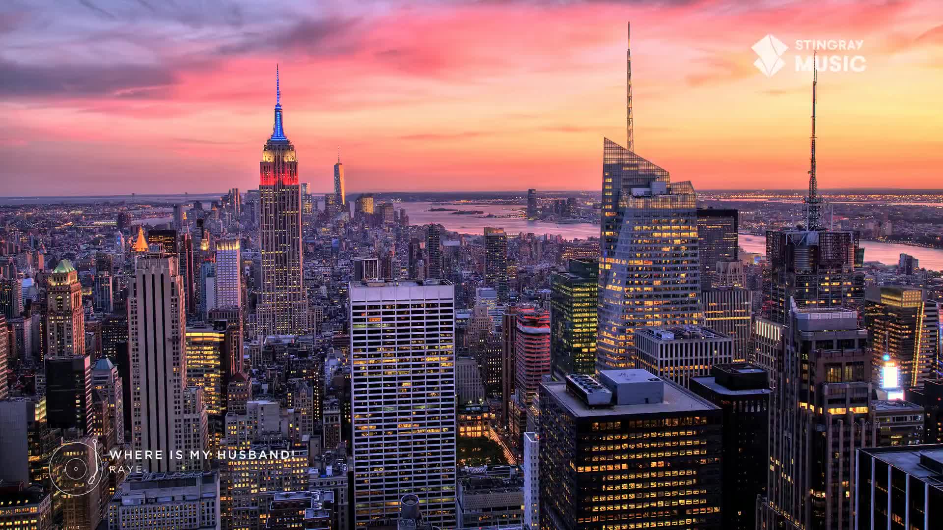 The Empire State Building glows red and blue against a pink and orange sky. Lights twinkle on in the surrounding skyscrapers as the sun sets over the water.
