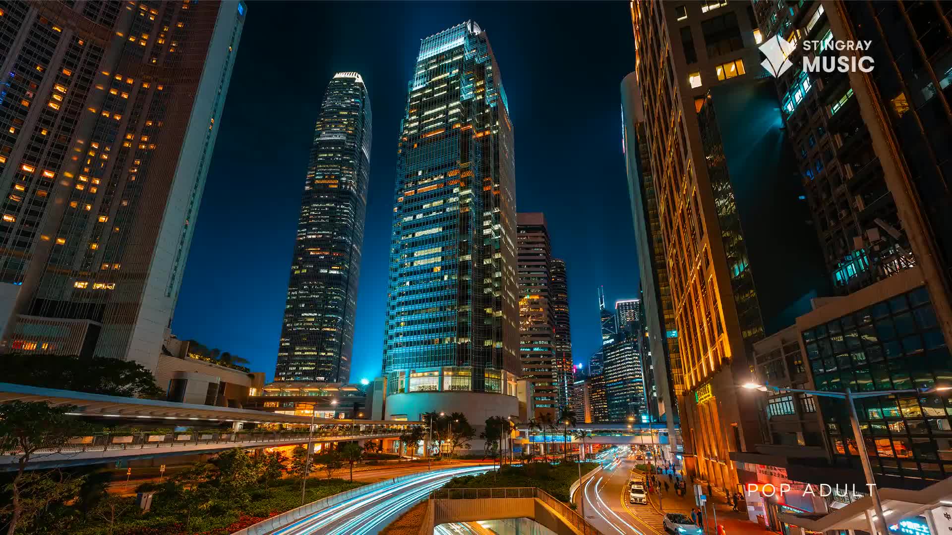 Headlights streak along the highway below, a river of light flowing between towering buildings. The night sky is a deep blue, punctuated by the warm glow of countless windows.