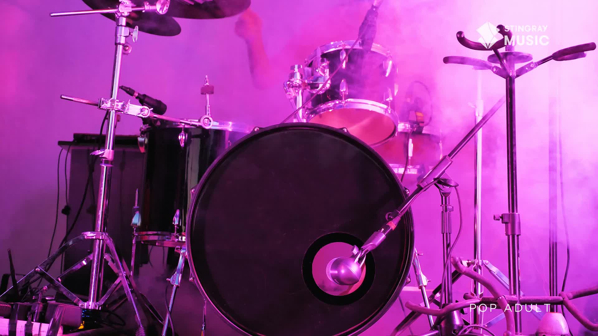 A drummer's arm is a blur as it strikes the snare, sending pink smoke swirling around the kit. The Stingray Music stage is bathed in a vibrant purple light, hinting at a lively pop adult performance.