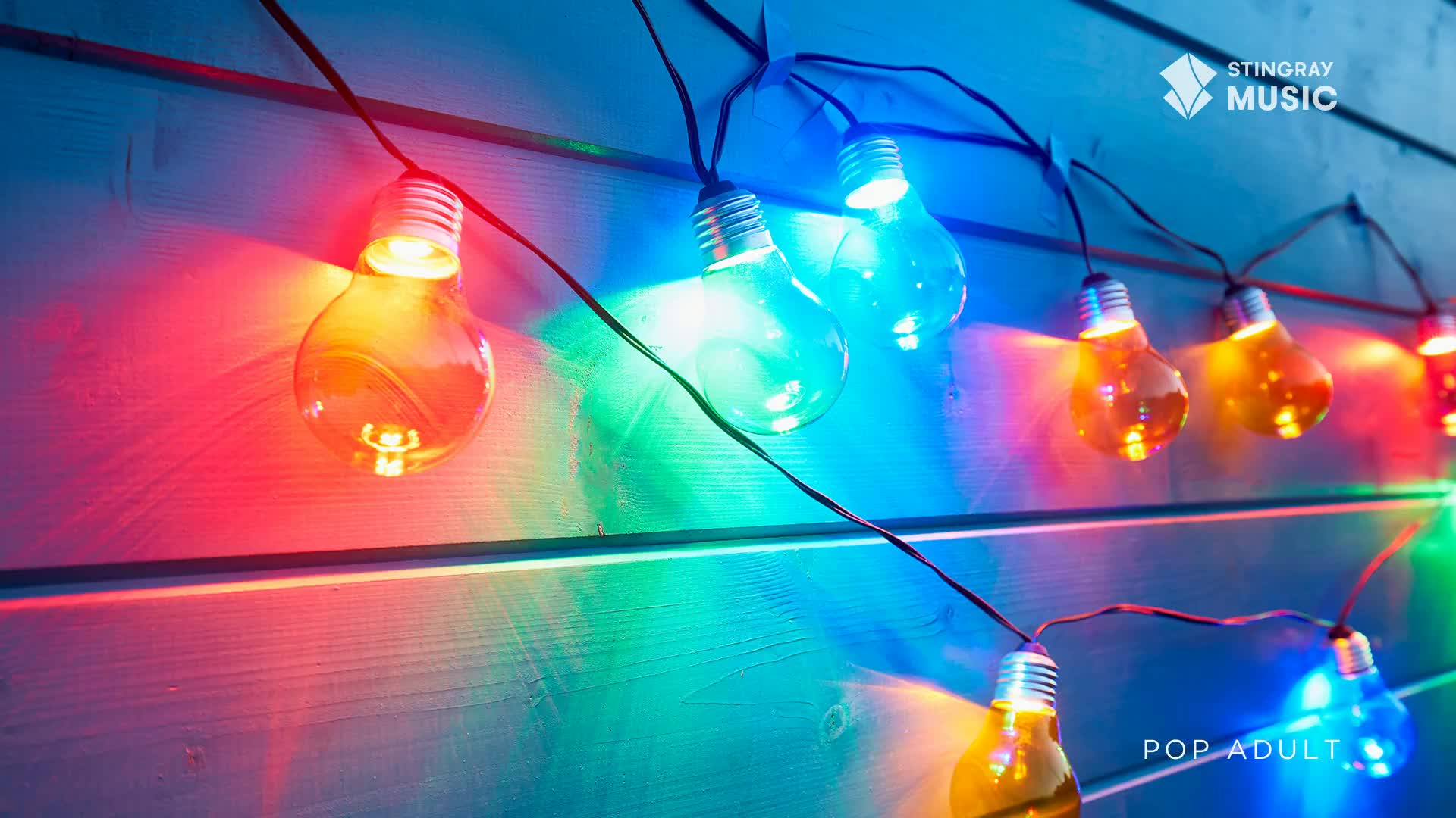 A string of clear bulbs glows with vibrant red, blue, and orange light against a wooden wall. These lights cast colourful shadows, creating a festive atmosphere.