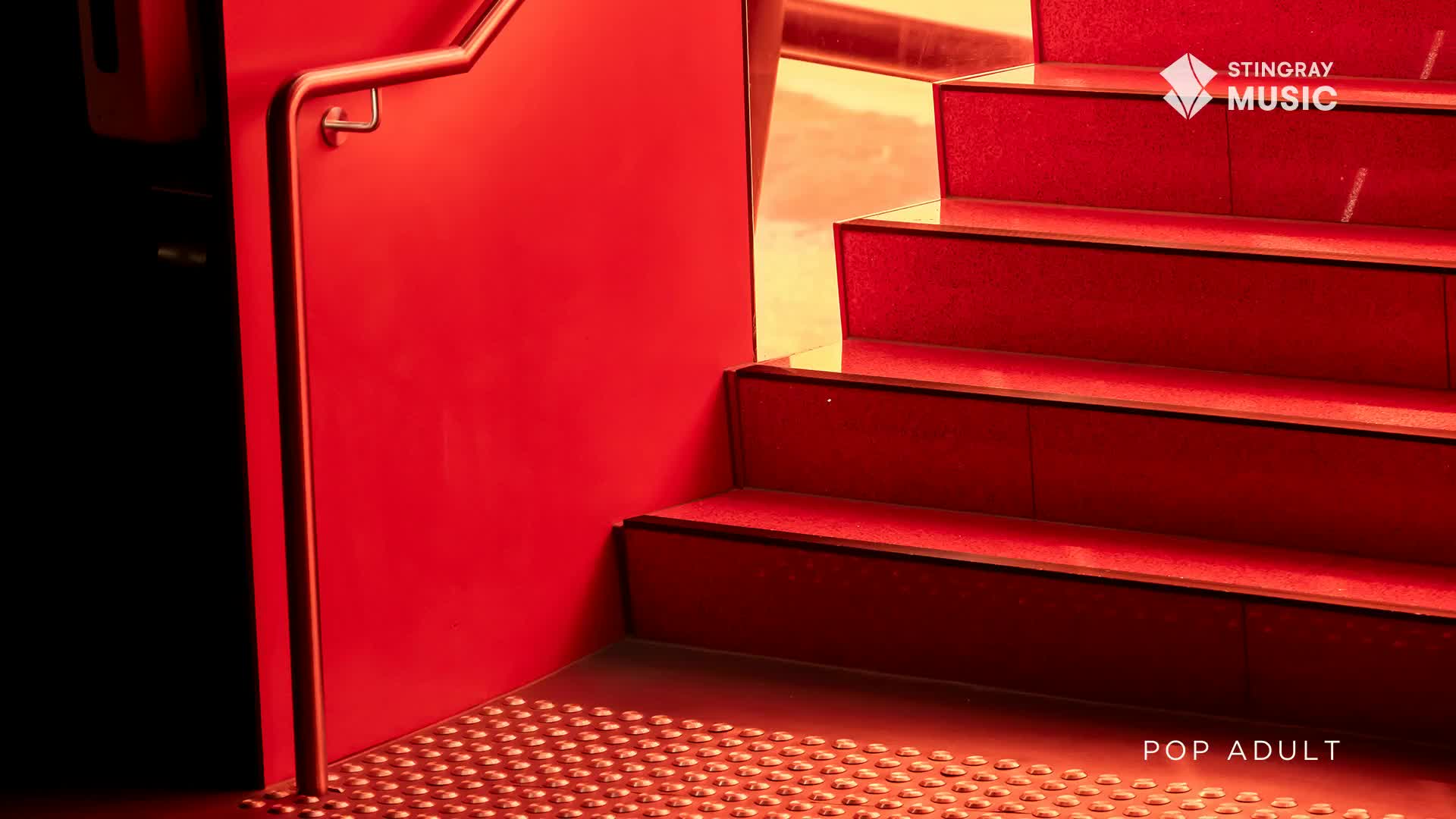 The red carpeted stairs ascend into a warm, golden light. A polished metal railing curves along the bright red wall.