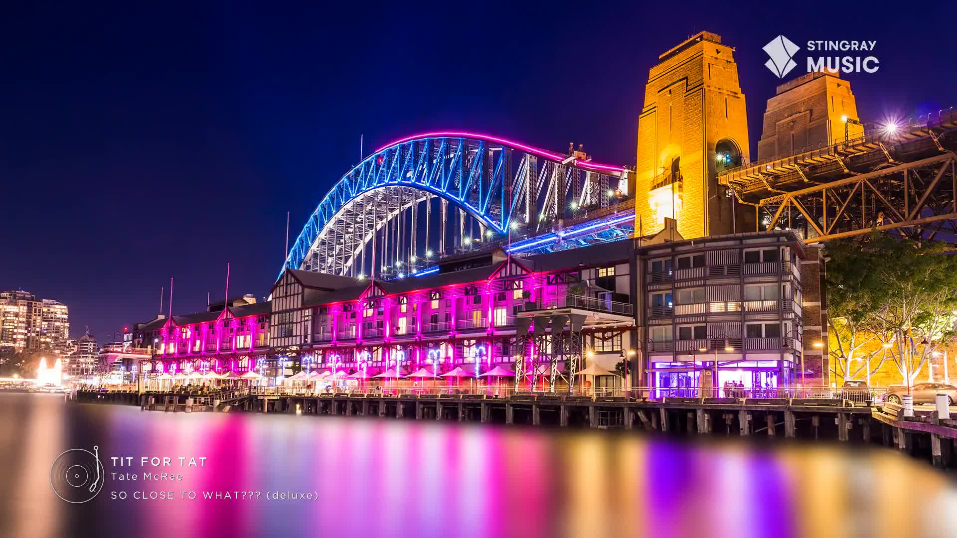 The Harbour Bridge glows with blue and pink lights, reflecting on the water. Below it, a row of buildings pulses with vibrant pink and purple illumination.