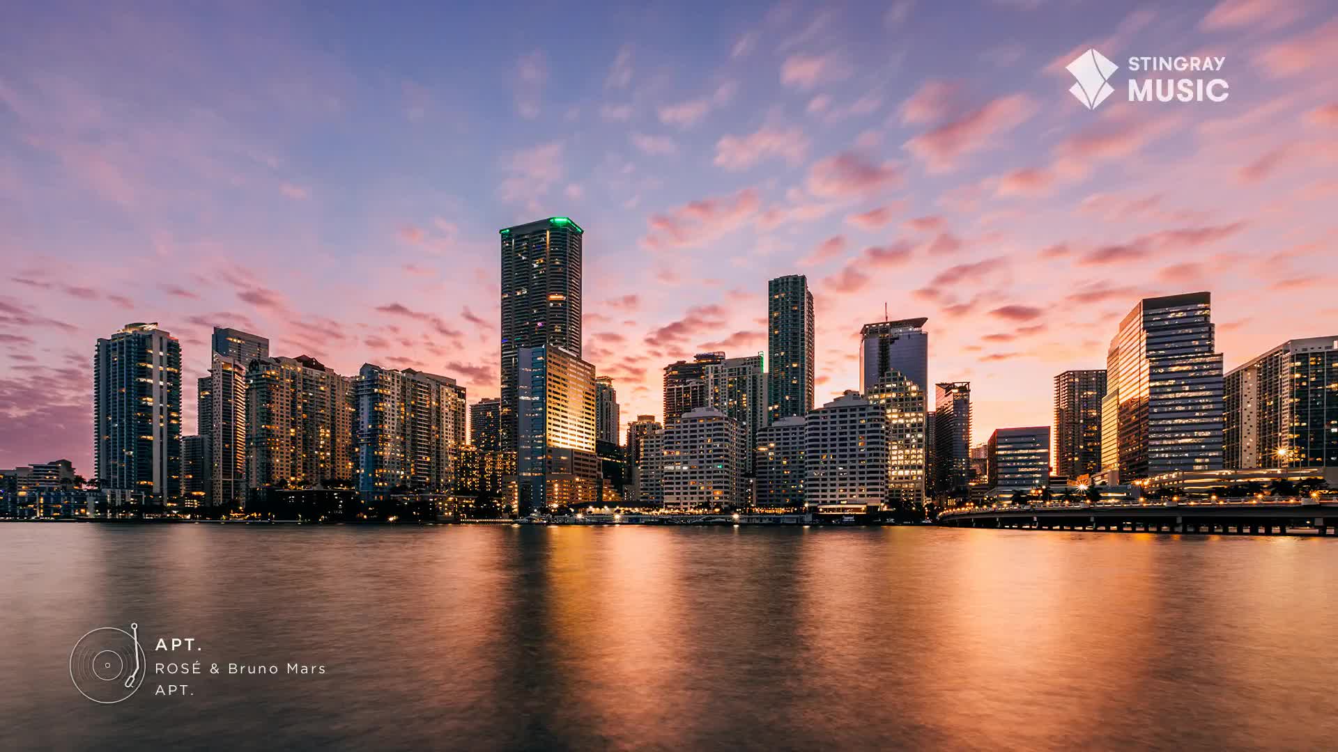 The sky above the city is painted with soft pink and orange hues as the sun sets. Lights begin to twinkle on in the towering buildings, reflecting on the calm water.