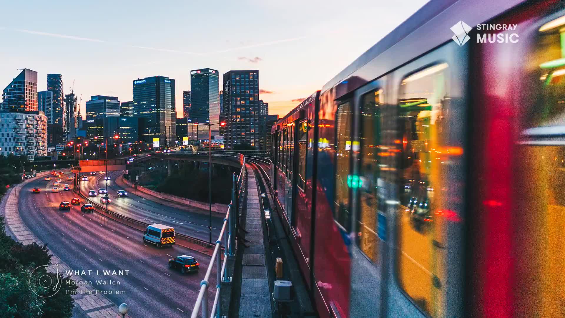 A red train rushes past on elevated tracks, its windows reflecting the city lights. Below, cars navigate a highway as dusk settles over the urban skyline.