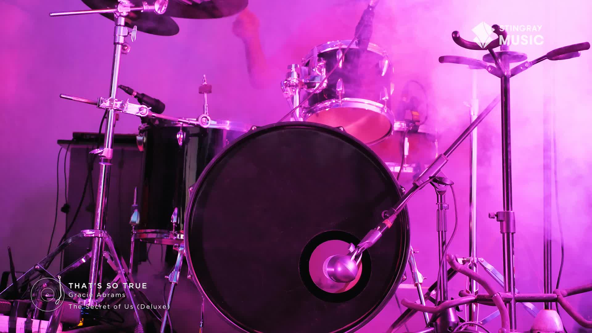 A drumstick is a blur above the snare, mid-beat. Pink stage lights cut through the haze surrounding the kit.