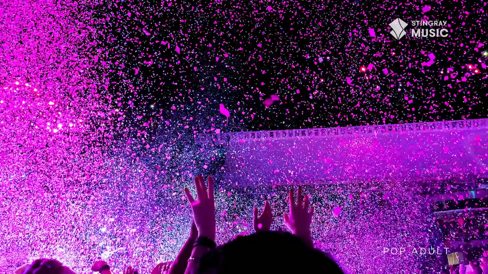 Confetti rains down in shades of pink and purple over a cheering crowd in Canada. Hands reach up towards the spectacle, bathed in the vibrant glow.
