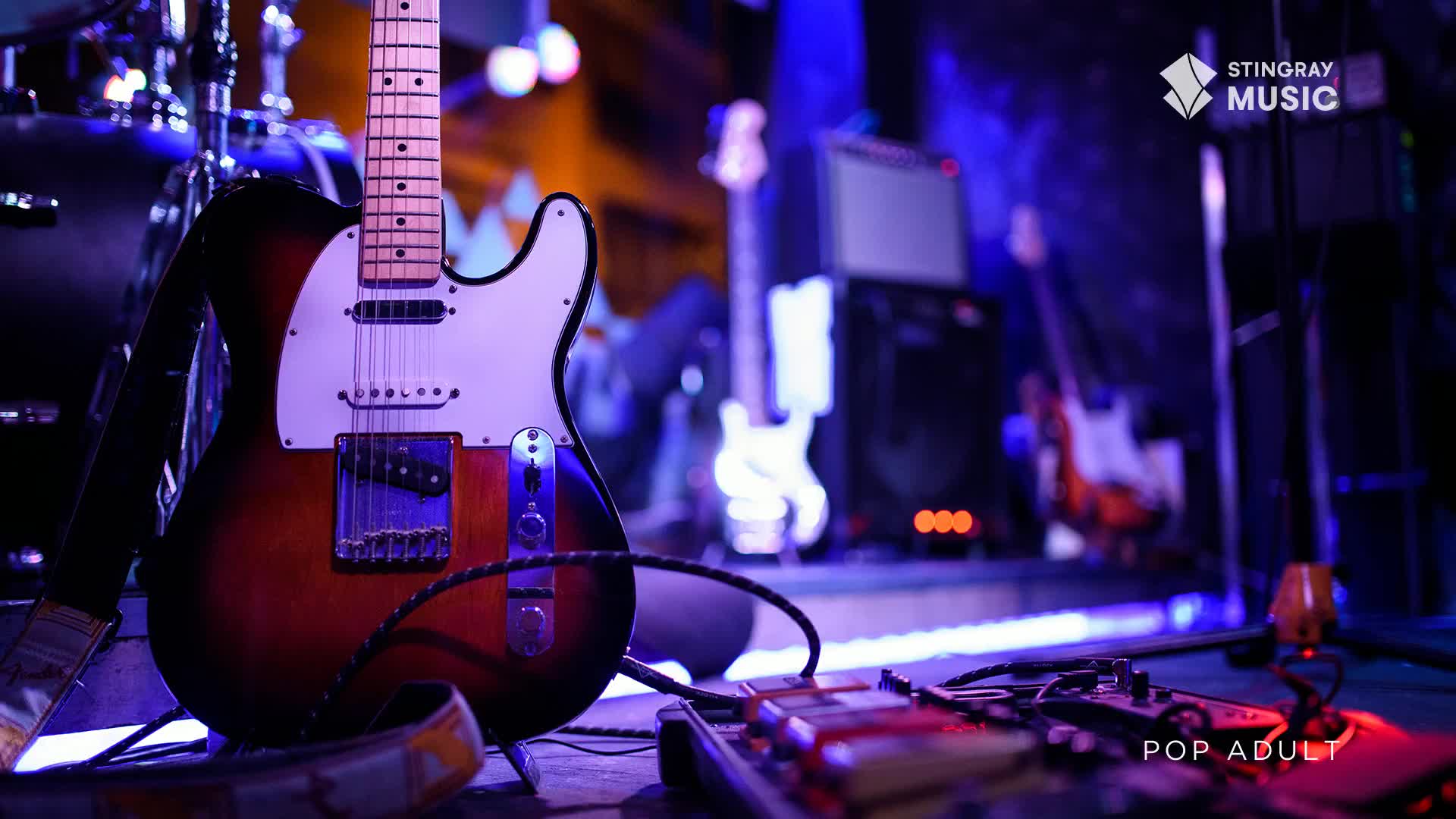 A sunburst electric guitar stands ready on a stage, its polished wood gleaming under the blue lights. Other instruments and amps are visible in the background, hinting at a live performance about to begin.