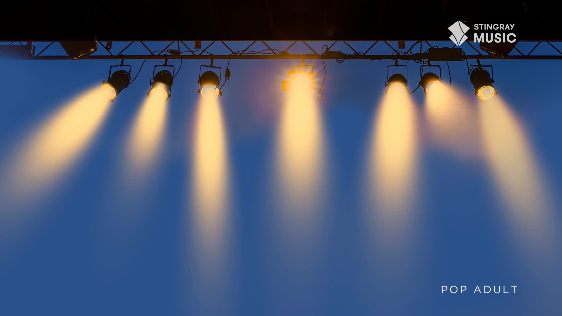 Bright spotlights cut through the evening sky, illuminating the stage for a Stingray Pop Adult performance. The beams of light, a warm golden hue, stretch downwards against the deep blue backdrop.