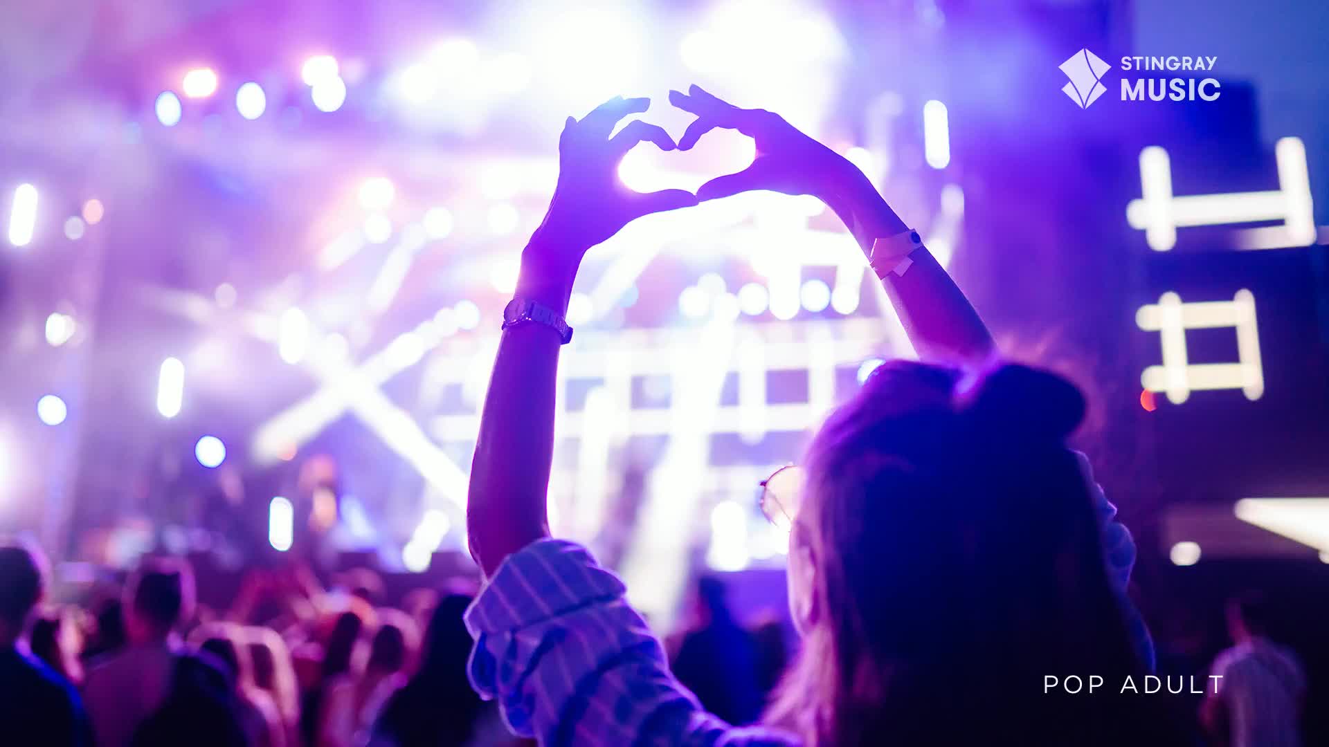 The stage lights pulse with purple and white as a fan holds their hands up, forming a heart. A crowd is gathered, enjoying the music.