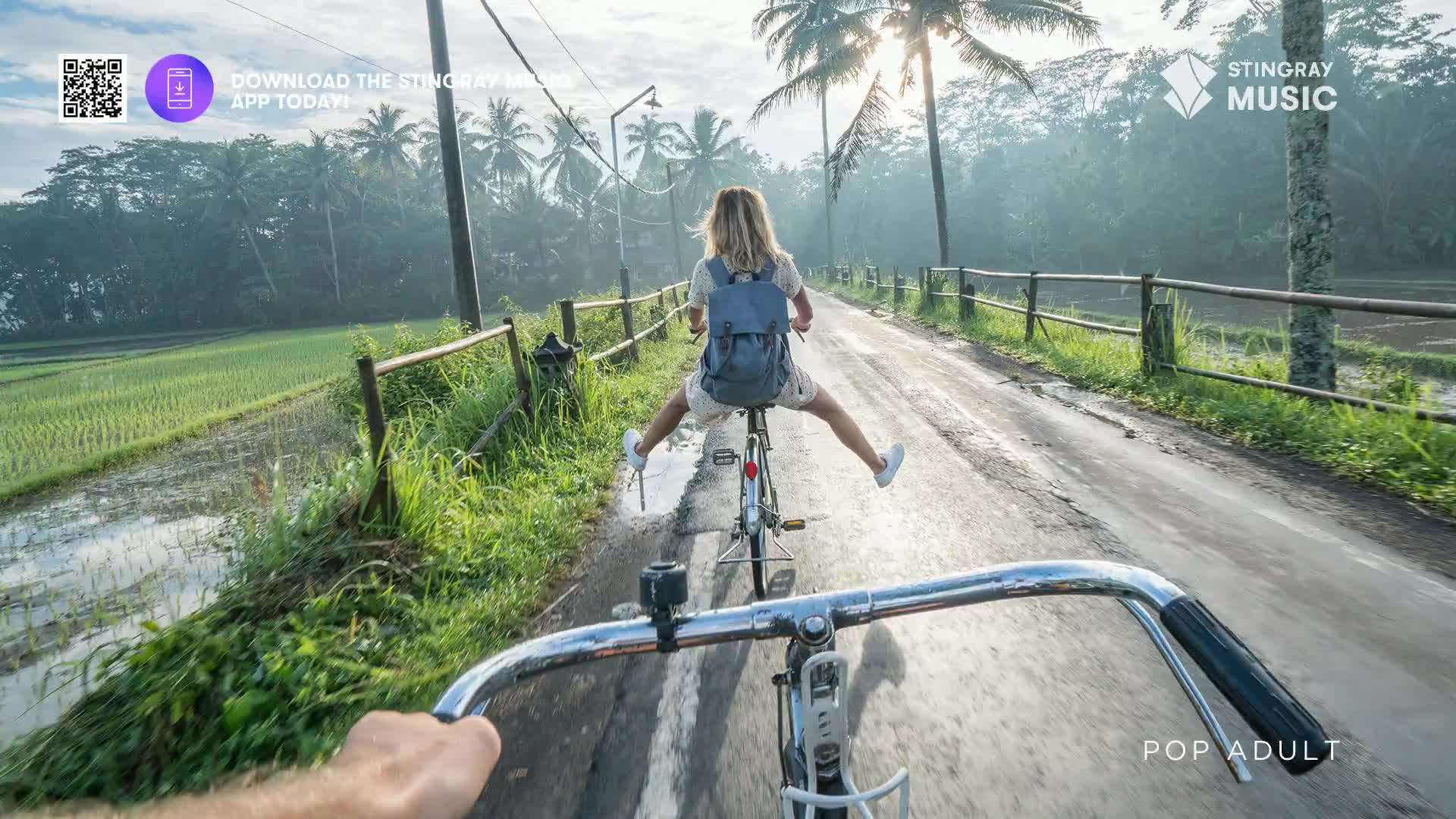 A woman on a bicycle rides ahead of me, her legs splayed wide as she pedals down a wet, rural road. Lush green rice paddies flank the path, and palm trees line the horizon under a bright, hazy sky. A woman on a bicycle rides ahead of me, her legs splayed wide as she pedals down a wet, rural road. Lush green rice paddies flank the path, and palm trees line the horizon under a bright, hazy sky.