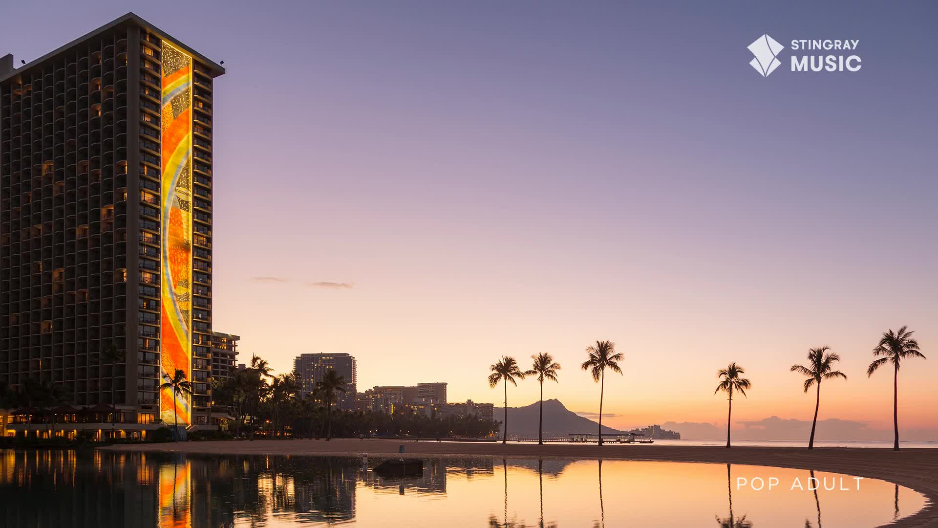 The sun is setting behind Diamond Head, casting a warm glow over the palm trees lining the beach. A tall building on the left is illuminated with a vibrant, abstract design. The sun is setting behind Diamond Head, casting a warm glow over the palm trees lining the beach. A tall building on the left is illuminated with a vibrant, abstract design.