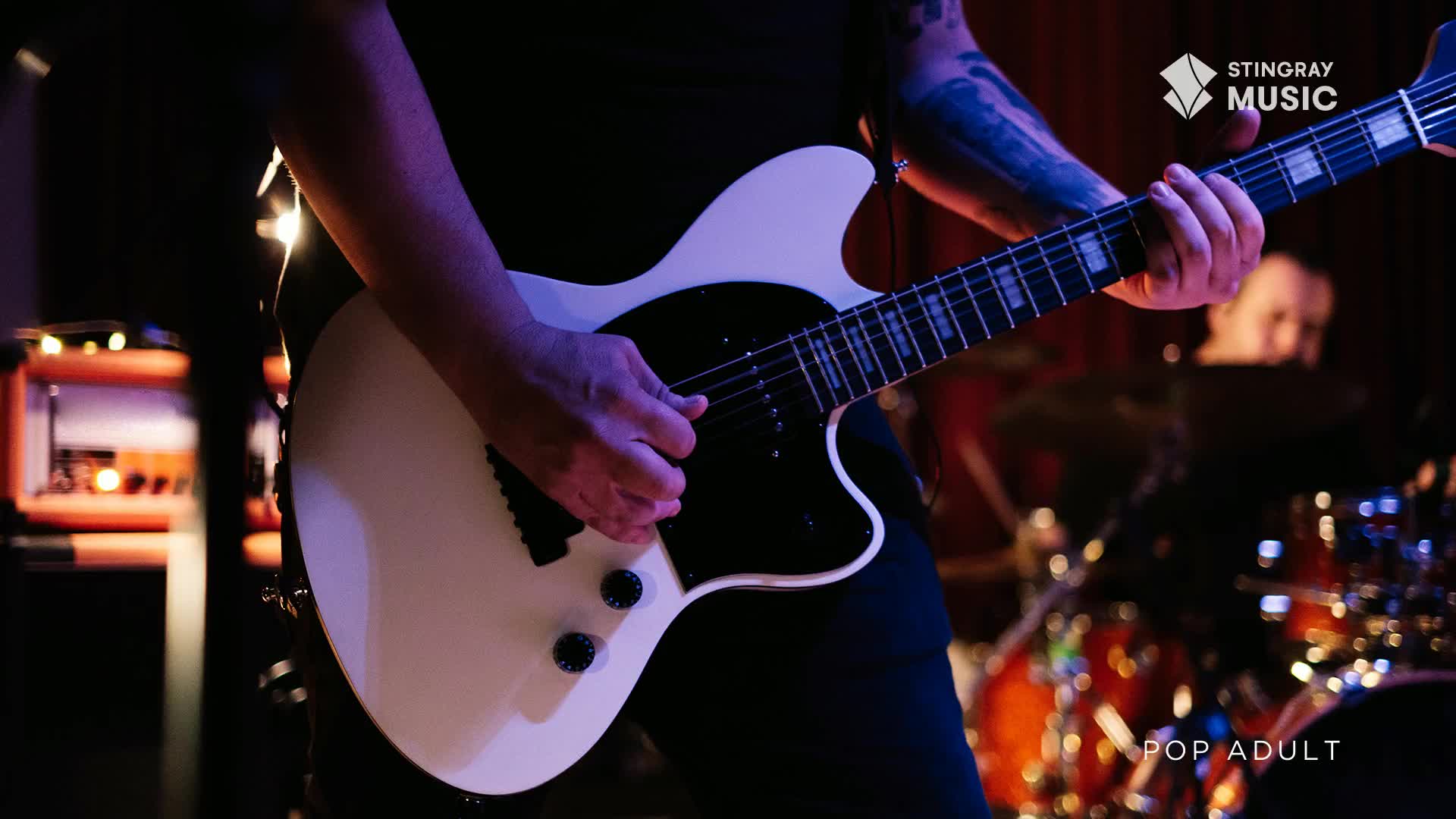 Fingers dance across the fretboard of a white electric guitar. A drummer's silhouette is visible behind the guitarist, bathed in warm stage lights.