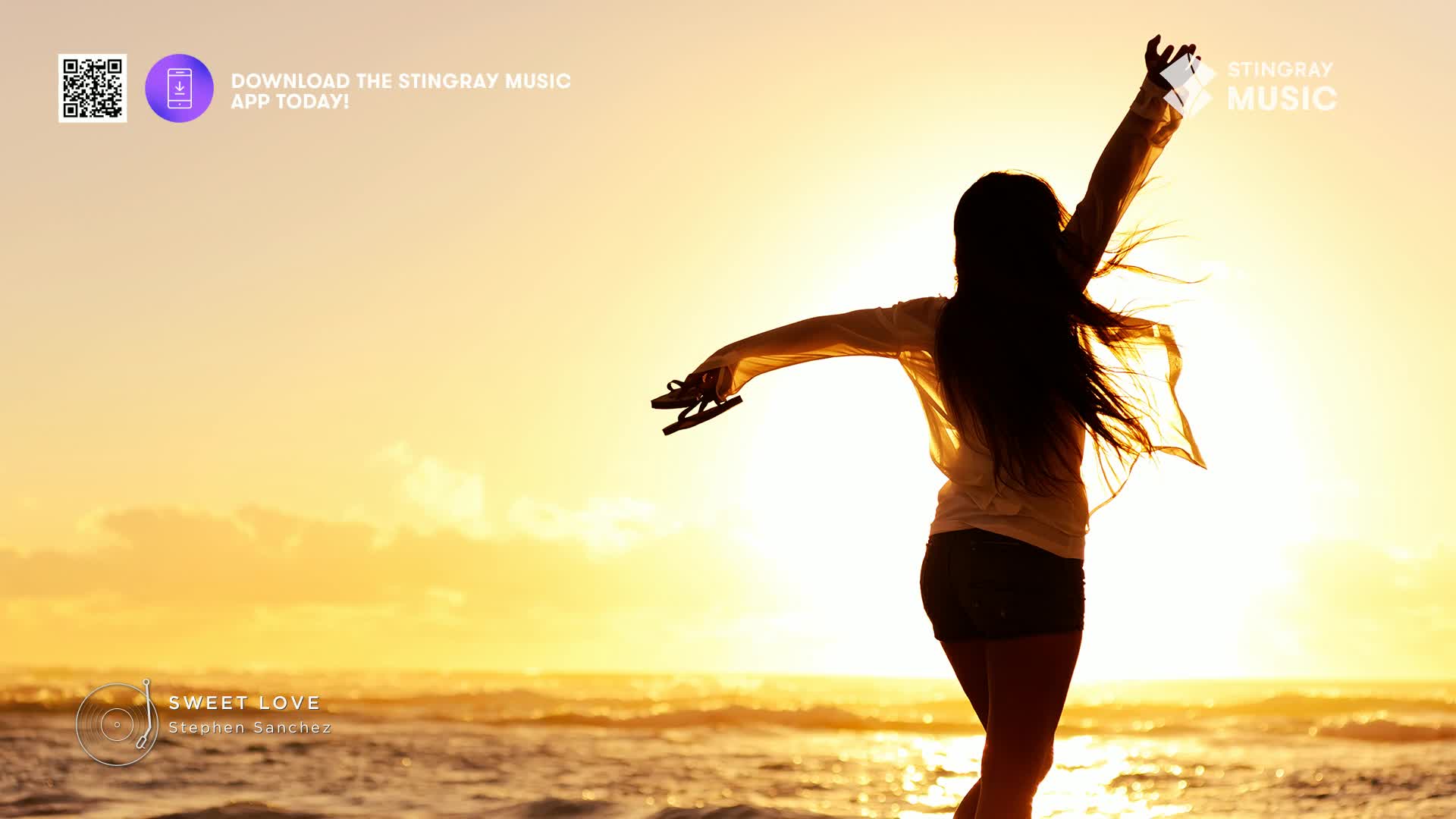 The sun dips low over the ocean, casting a warm glow as a woman with long, dark hair dances on the shore. She holds her flip-flops aloft, her arms outstretched in a gesture of pure joy.