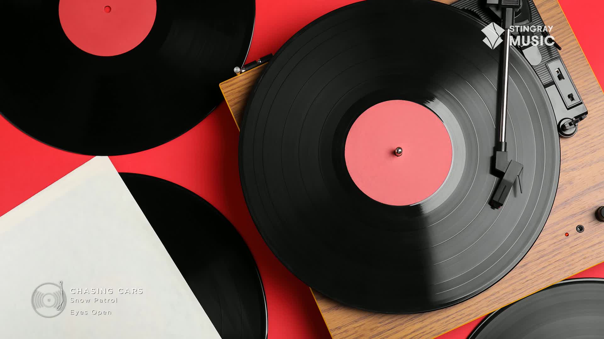 A needle rests on a spinning vinyl record, its pink label catching the light. Nearby, another record sits in its sleeve, the title "Chasing Cars" by Snow Patrol visible.