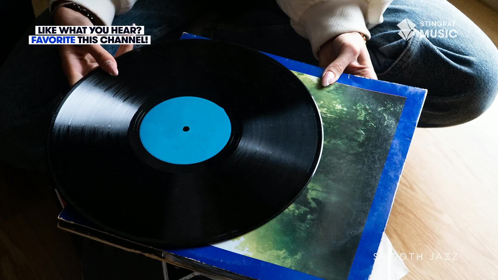 A person's hands hold a shiny black vinyl record, its blue label catching the light. The album cover beneath it hints at a smooth jazz vibe, perhaps something from Canada's Stingray Music.
