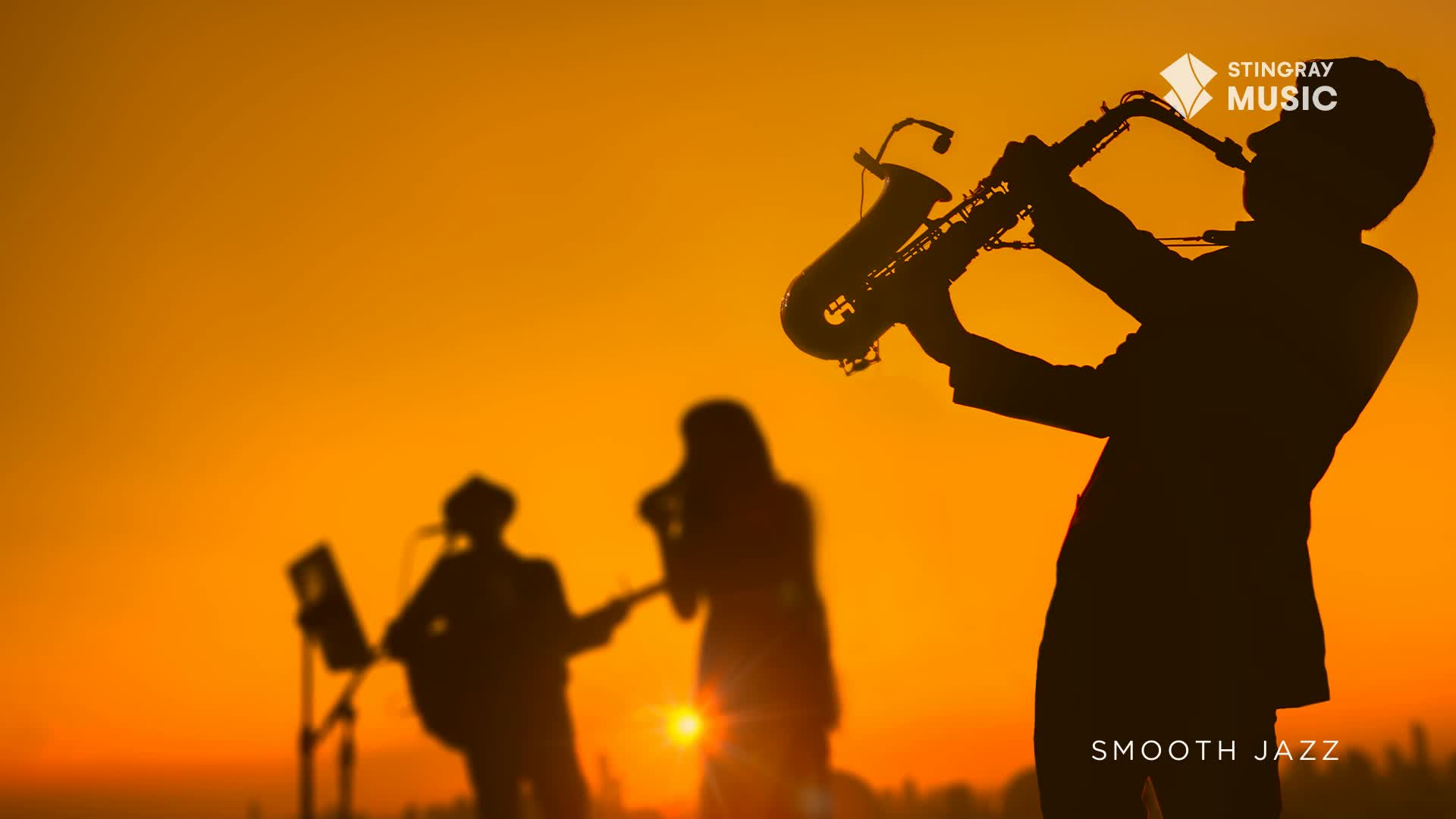 The silhouette of a musician blows into a saxophone, its shape defined against an orange sky. A band plays in the background, their forms hazy in the warm light of the setting sun.
