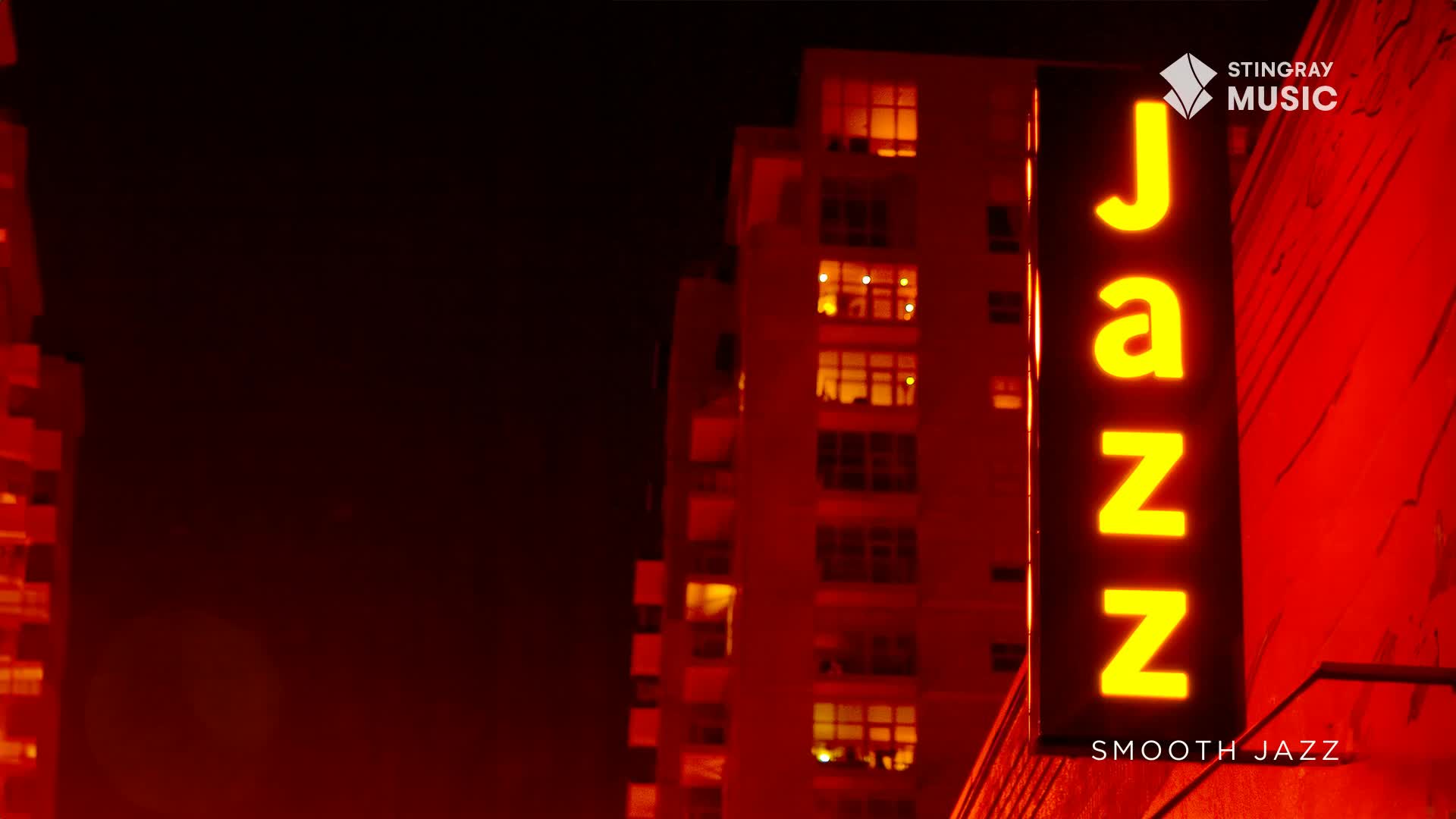 The neon sign glows, spelling out "Jazz" against the red-lit wall. Beyond, the windows of a building are lit, a warm contrast to the night.
