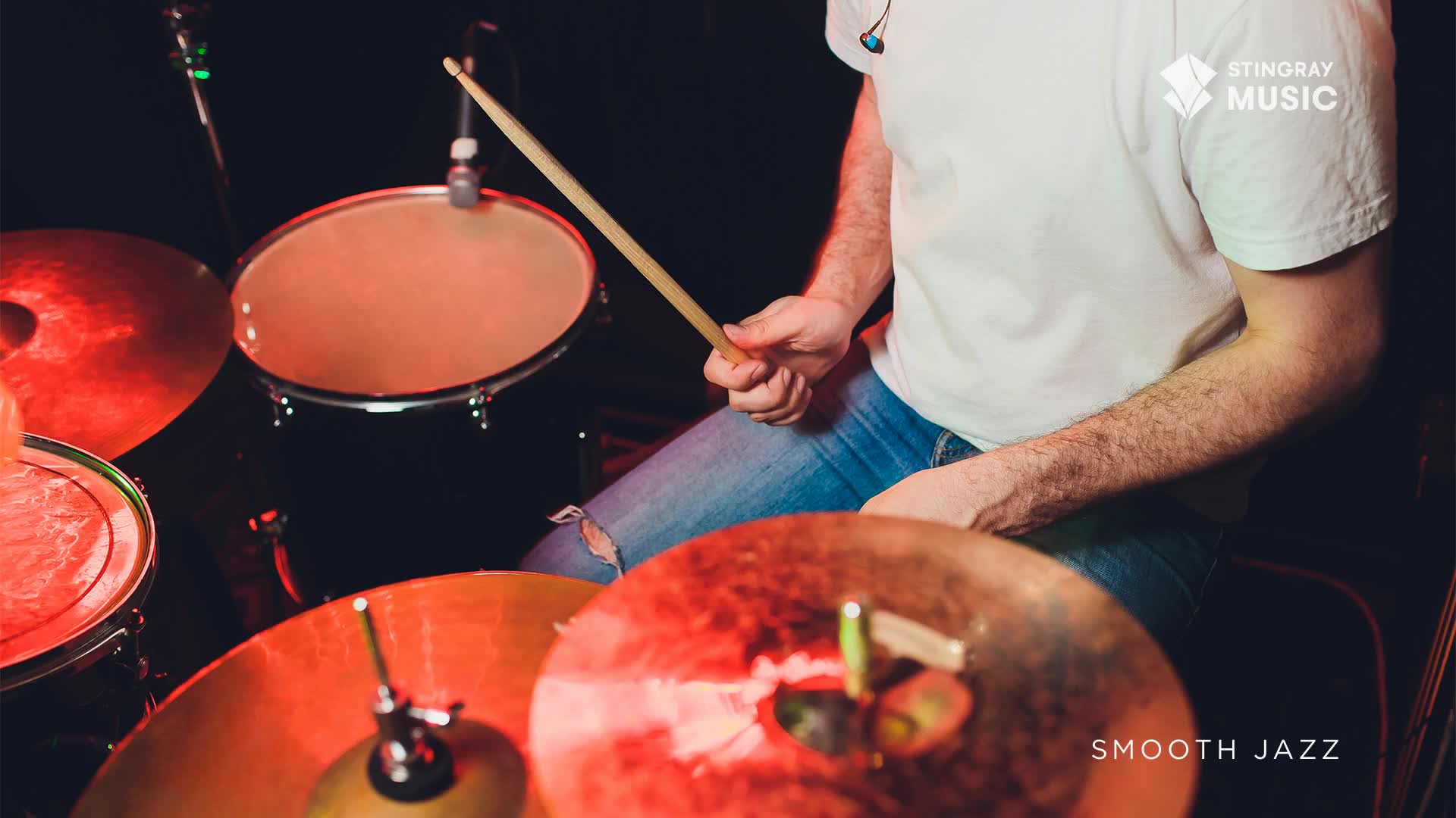 A drummer, bathed in red light, is poised over his kit. The wooden stick is about to strike the snare, ready to keep time with the Stingray Smooth Jazz.
