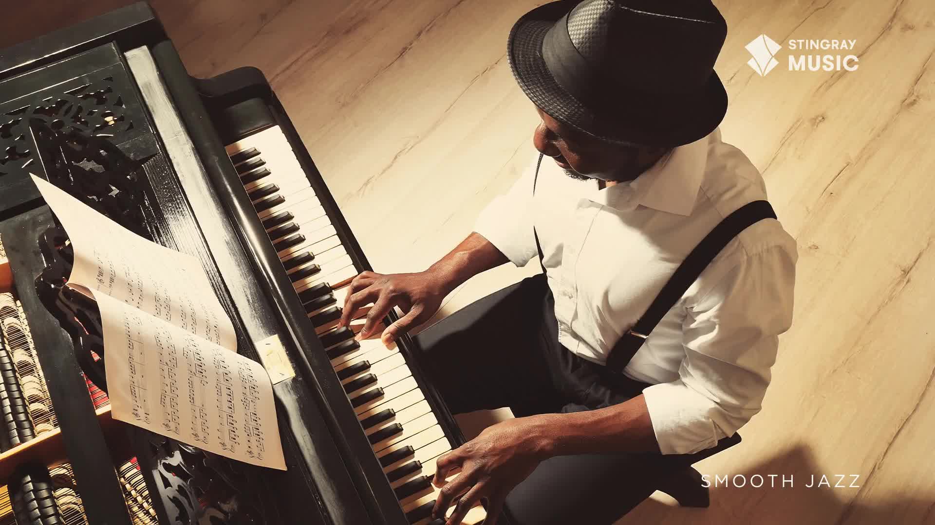 A man in a fedora and suspenders sits at a piano, his fingers dancing across the keys. Sheet music rests on the stand, while the Stingray Smooth Jazz logo is visible in the corner.
