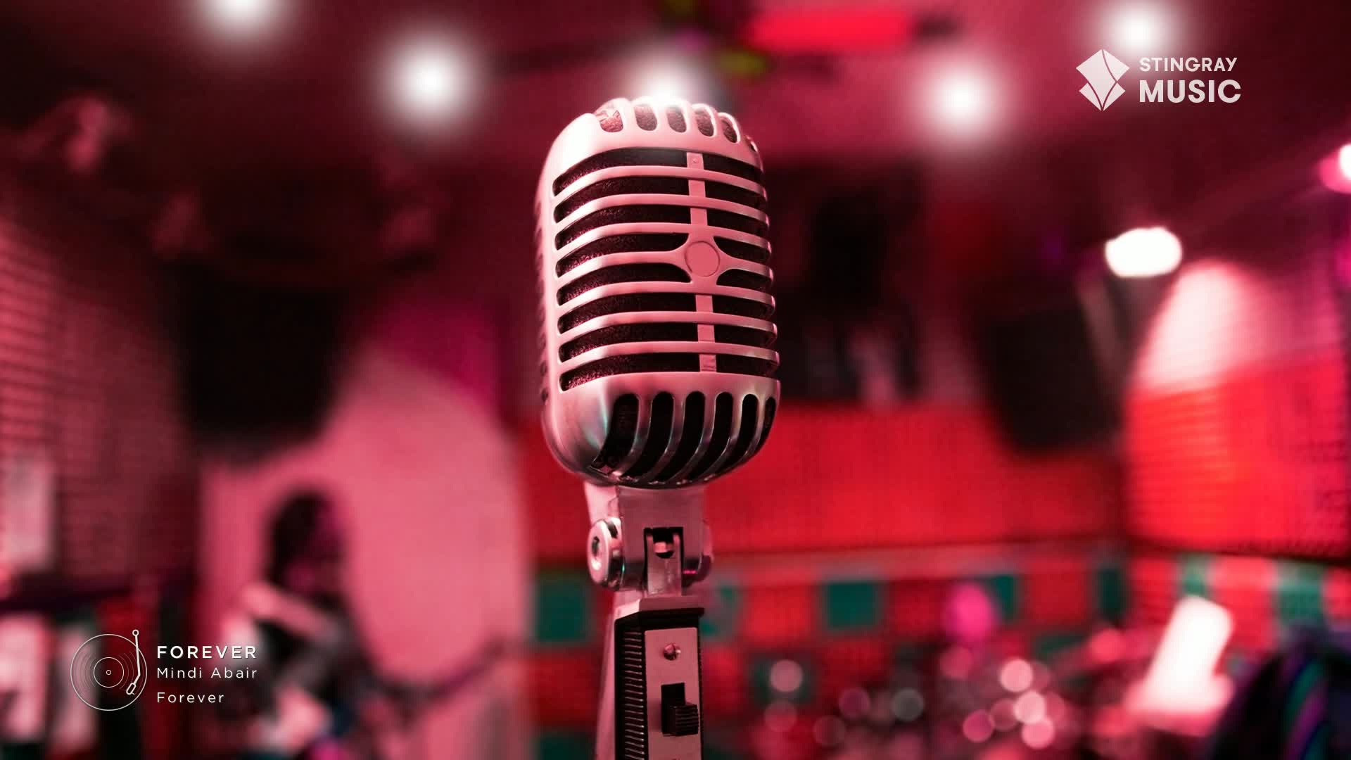 A vintage microphone stands center stage, bathed in the warm glow of red lights. In the background, a band is setting up, instruments ready for a Stingray Smooth Jazz performance.