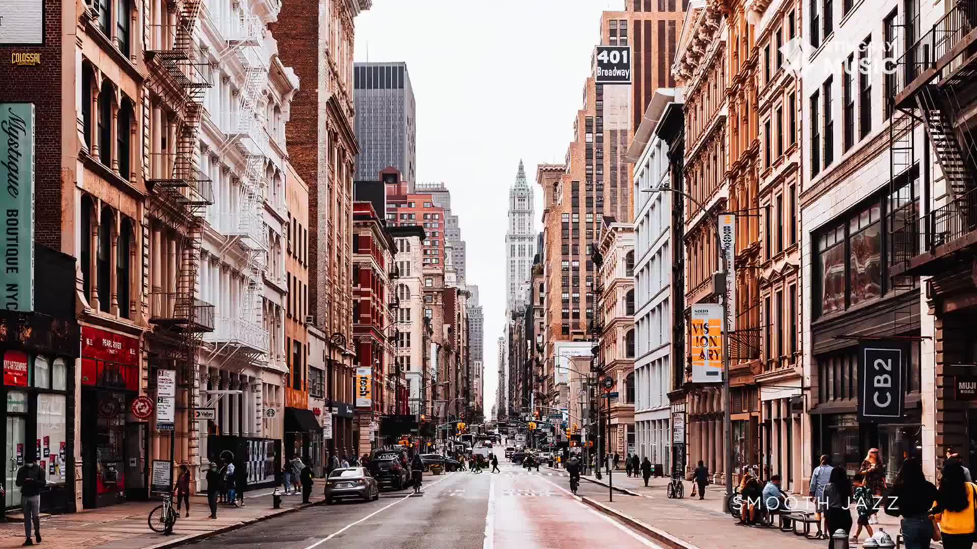 Cars are moving down a street lined with tall buildings. People stroll along the sidewalks, some pausing to chat.