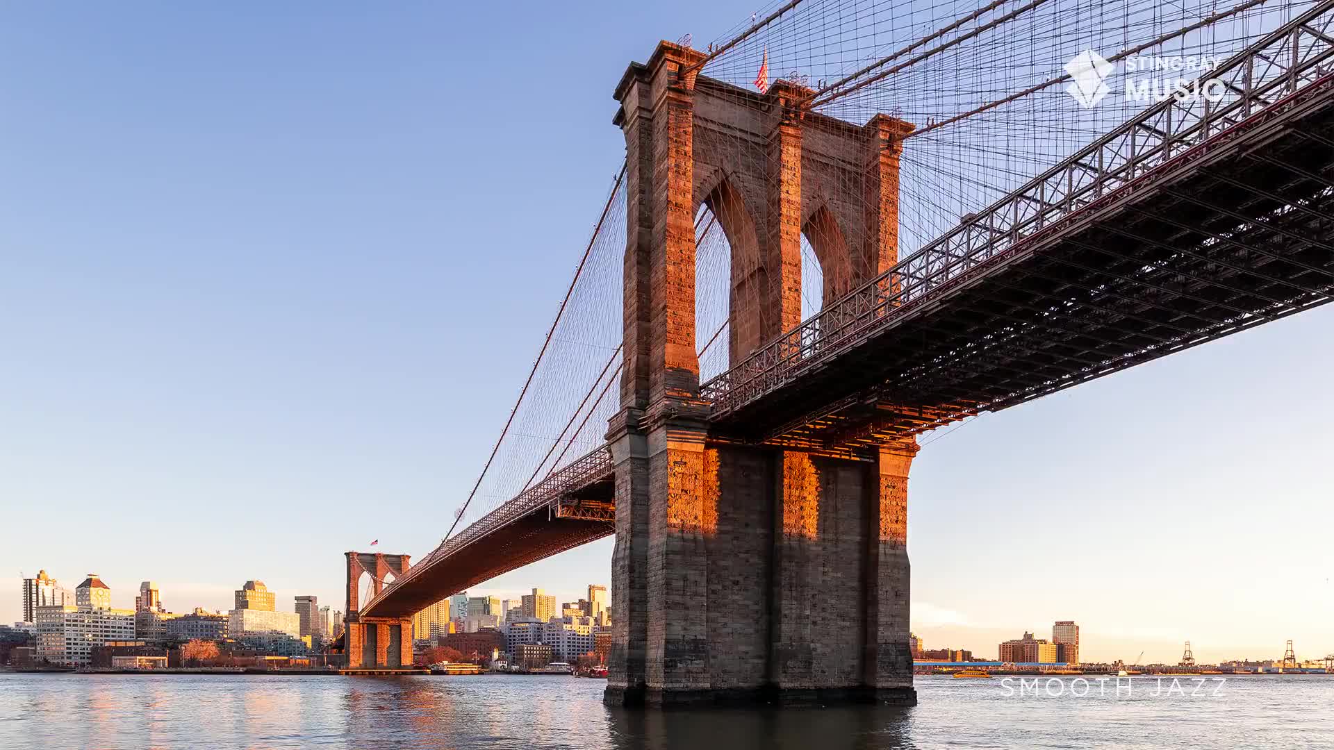 The sun casts a warm glow on the stone towers of the Brooklyn Bridge. Beyond, the city skyline stretches out under a clear blue sky, a perfect backdrop for some smooth jazz.