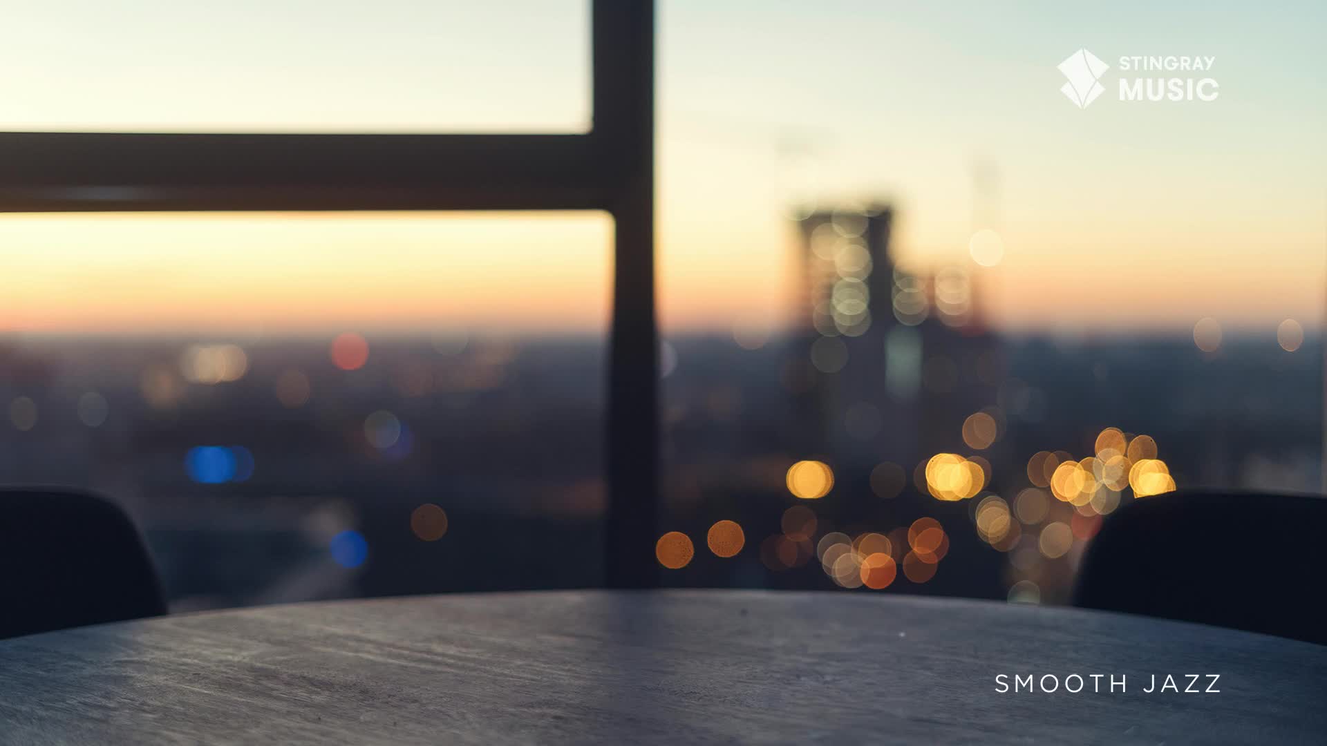 The city lights twinkle below as the sun dips below the horizon, painting the sky in soft oranges and yellows. A table sits in the foreground, ready for a quiet evening, perhaps with some Stingray Smooth Jazz playing.