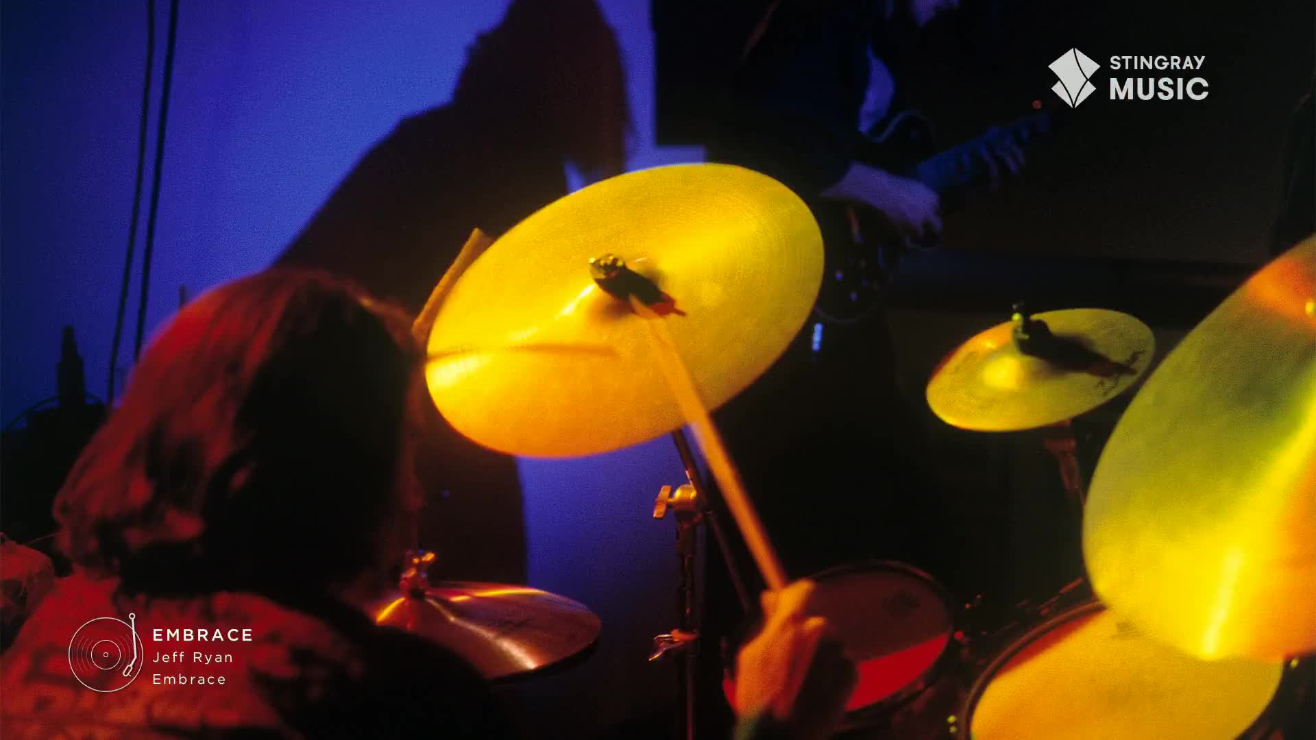 A drummer's sticks strike a bright cymbal, its surface catching the stage lights. The rhythmic beat accompanies a guitarist playing nearby, their instruments filling the air with smooth jazz.