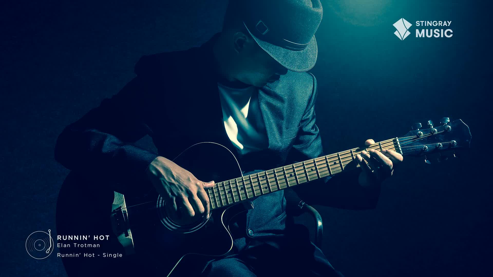 A man in a fedora and suit plays an acoustic guitar, his fingers moving across the fretboard. This is Elan Trotman's "Runnin' Hot," a single from Stingray Music.