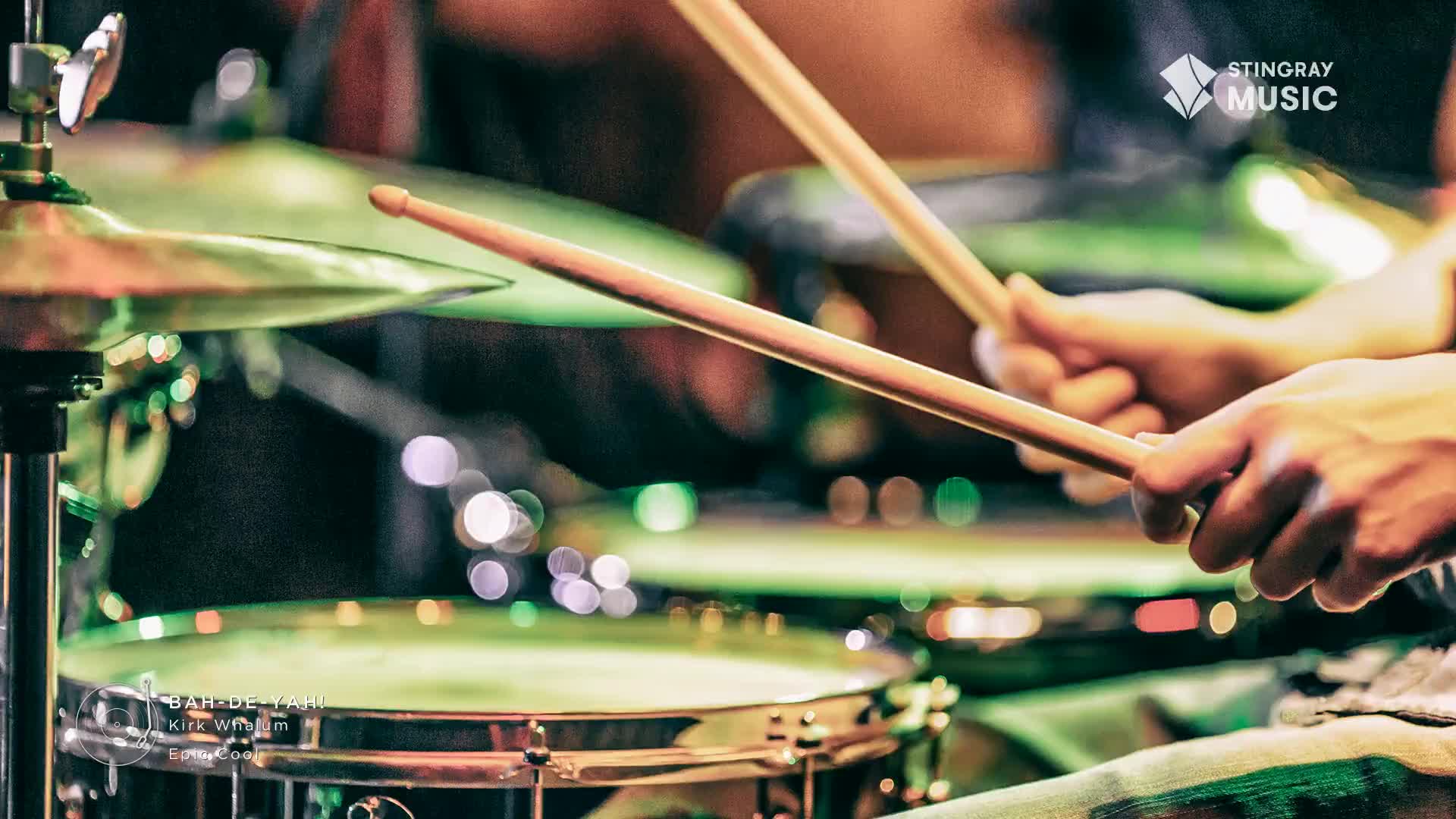 Drumsticks are poised above a snare drum, ready to strike. The lights behind the kit create a soft, colorful glow, suggesting a live performance for Stingray Smooth Jazz.