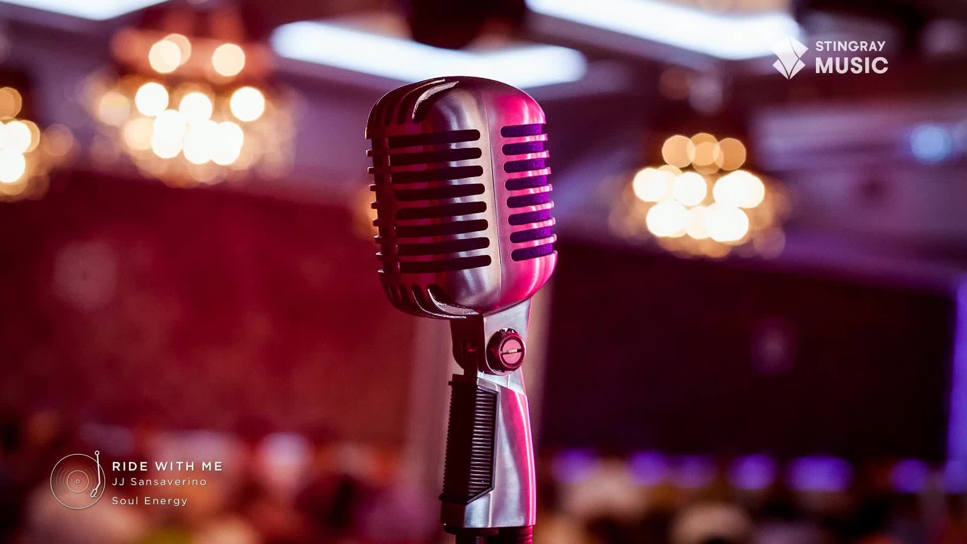 A classic chrome microphone stands center stage, bathed in a warm, red-tinted light. Behind it, the soft glow of chandeliers and purple stage lights hint at a lively venue, perhaps a jazz club in Canada.
