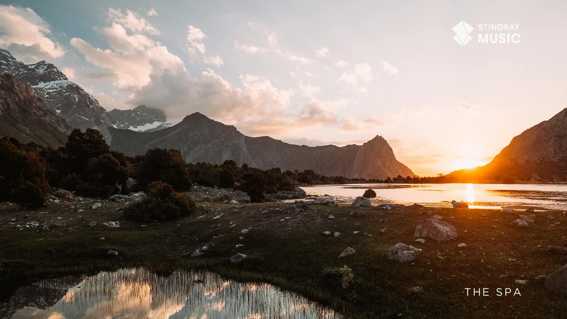 Sunlight spills across the water, reflecting the fiery sky and the imposing mountain range. The scene, branded by Stingray Music, evokes a tranquil spa experience in the heart of Canada's wilderness.
