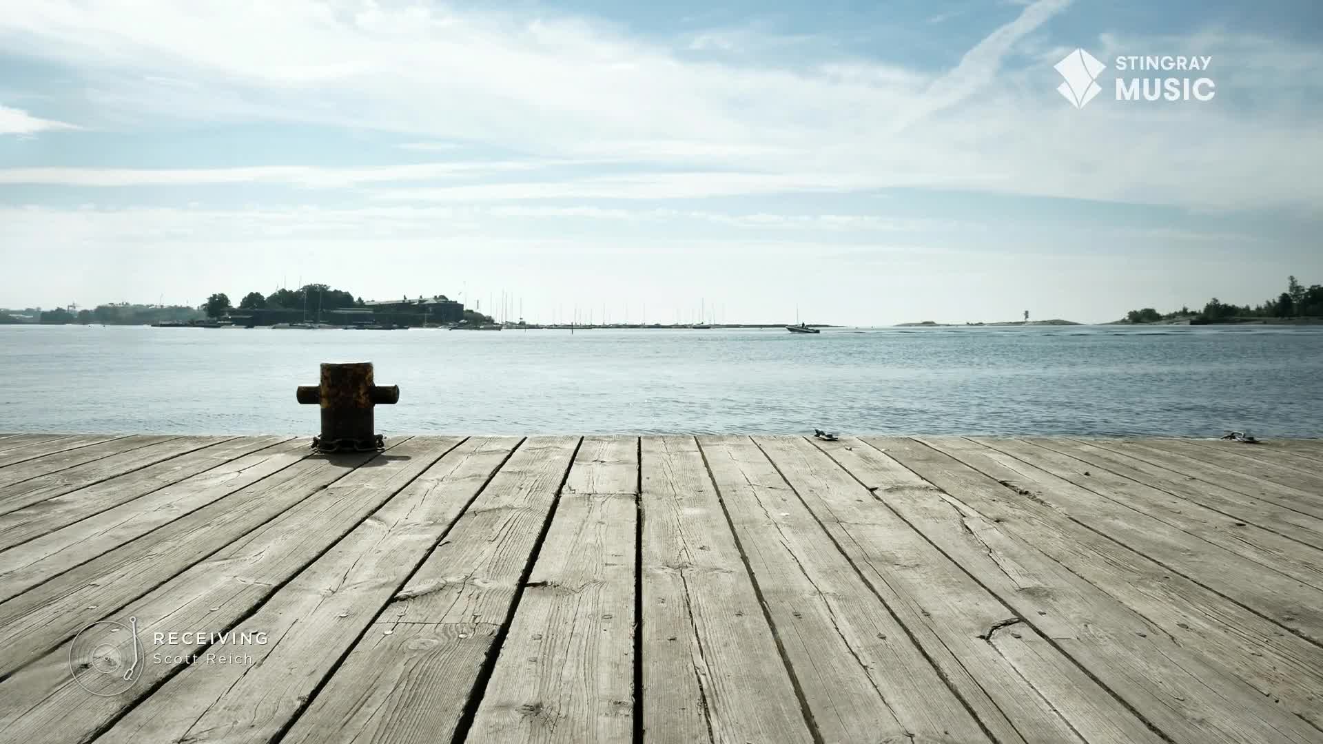 The weathered wooden dock stretches out toward the water, with a small boat puttering across the lake. A distant island sits on the horizon under a bright, cloud-streaked sky.
